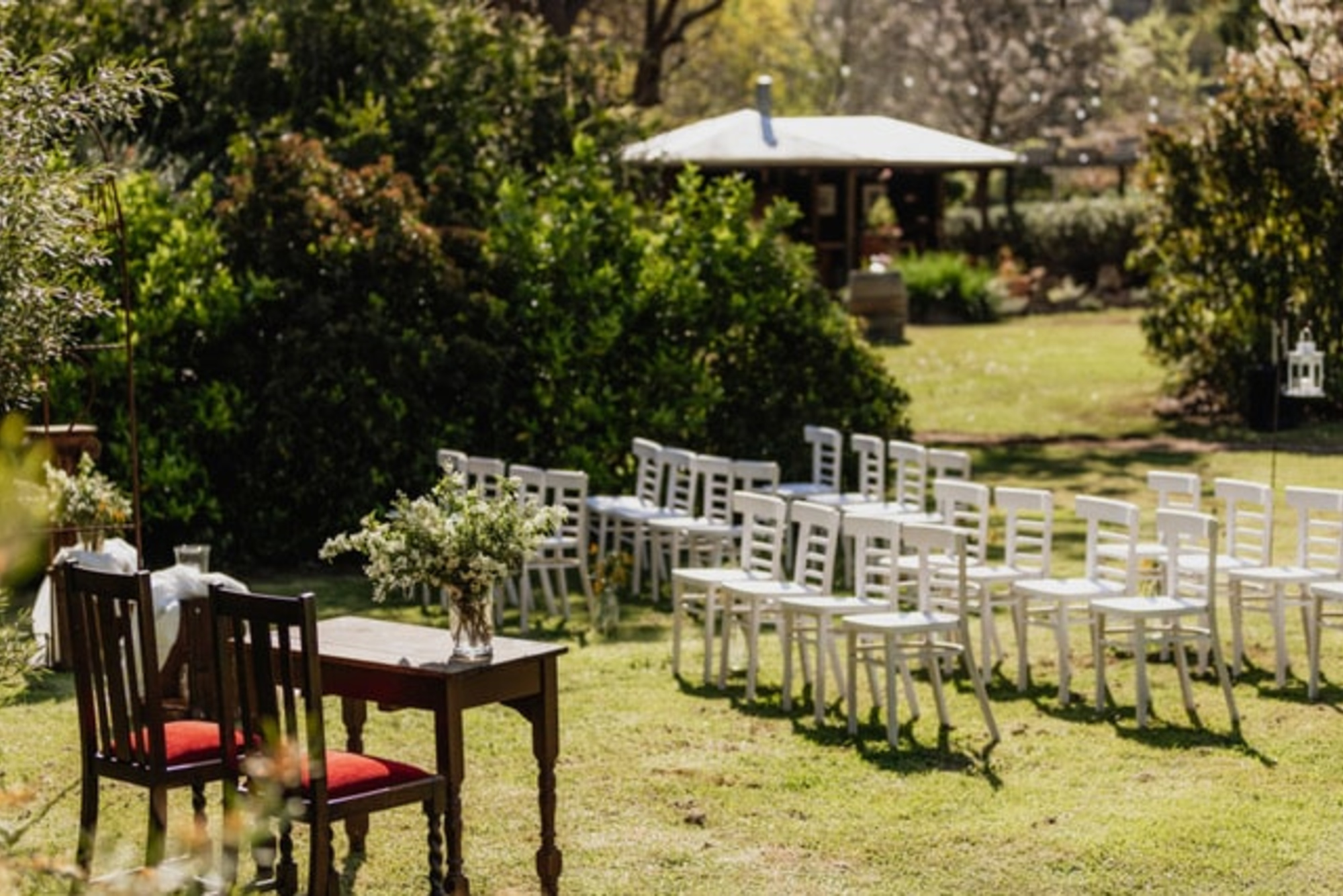 Outdoor garden wedding ceremony setup with white chairs and a wooden signing table on a sunny lawn.