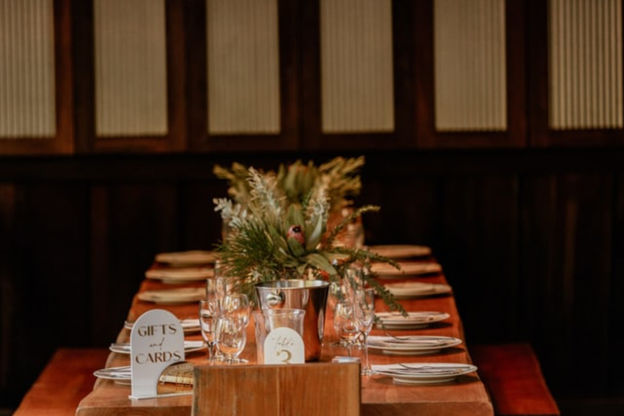 Warm rustic wedding reception table set with plates, glassware, and a greenery centerpiece in a cozy indoor venue.