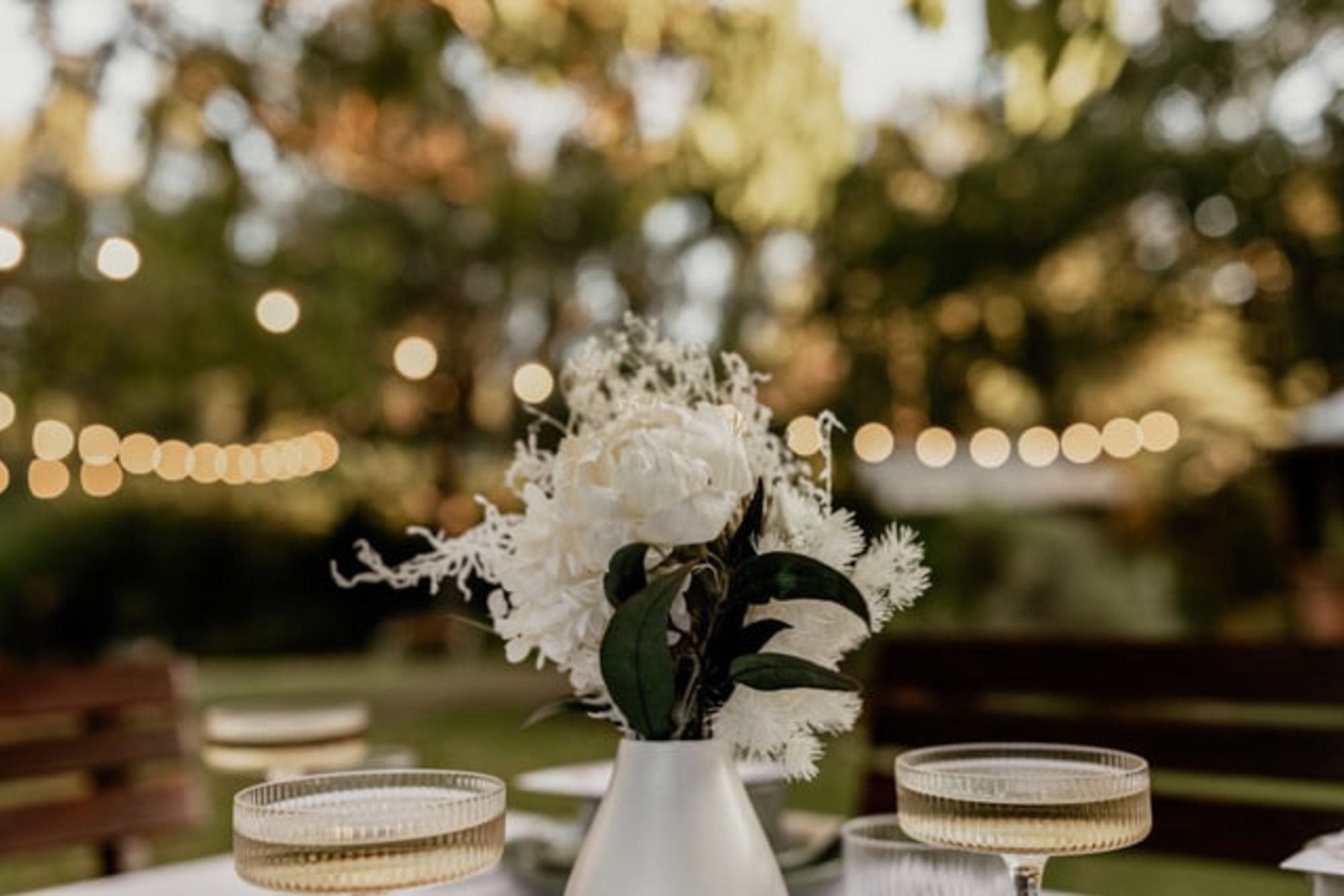Outdoor wedding table with a white floral centerpiece and string lights softly glowing in the background.