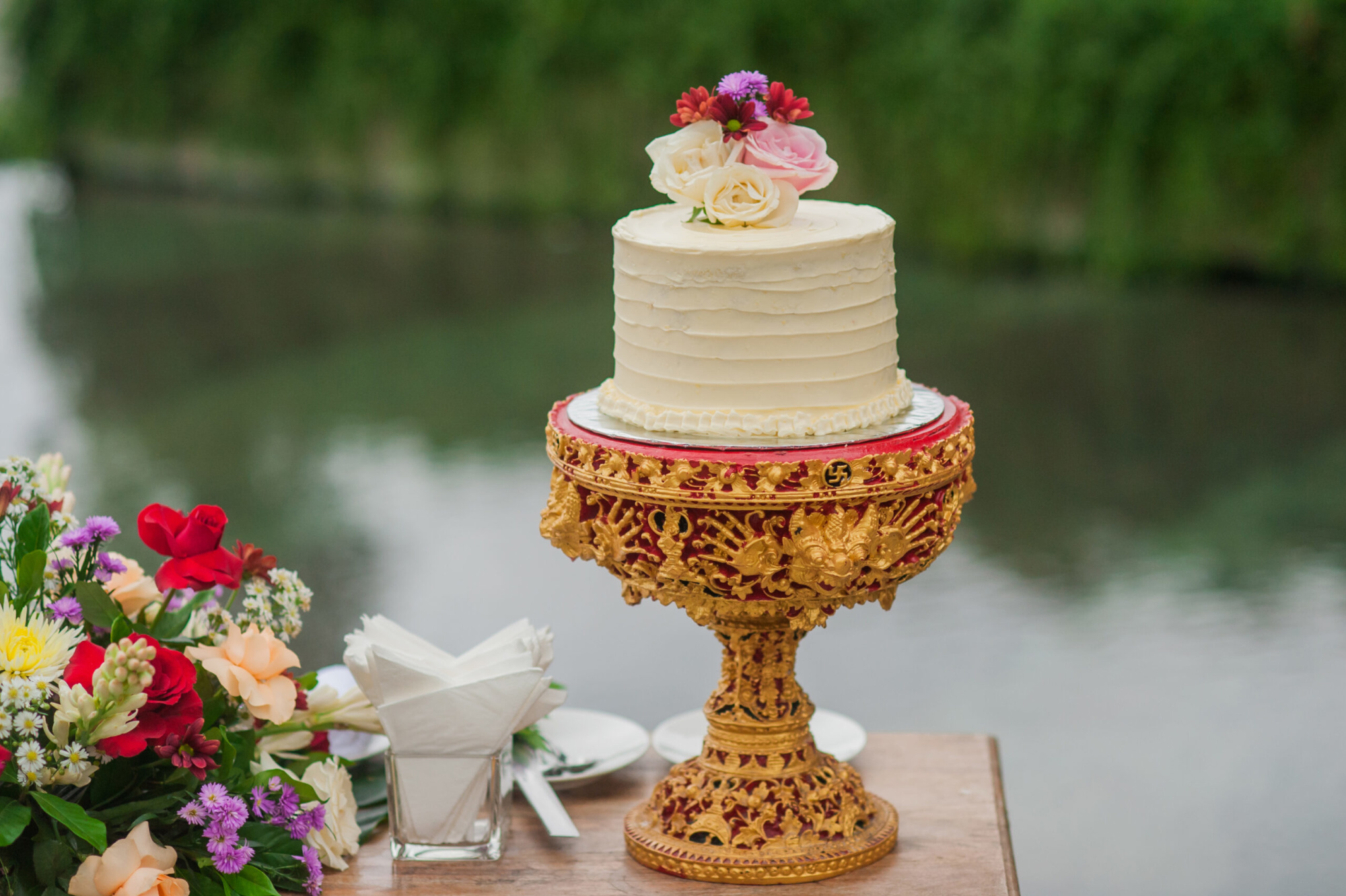 Single-tier white wedding cake with floral topper on ornate gold stand by a riverside table with colorful flowers.