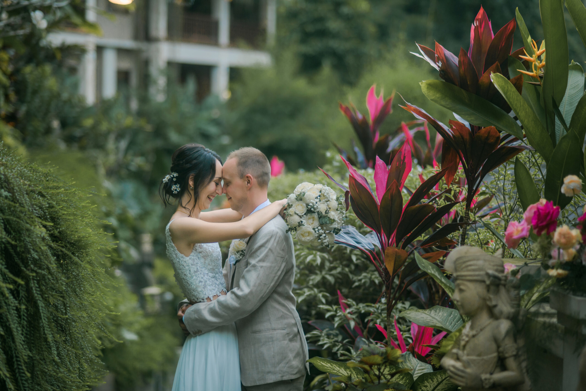 Bride and groom embrace in a lush tropical garden surrounded by vibrant flowers and greenery.