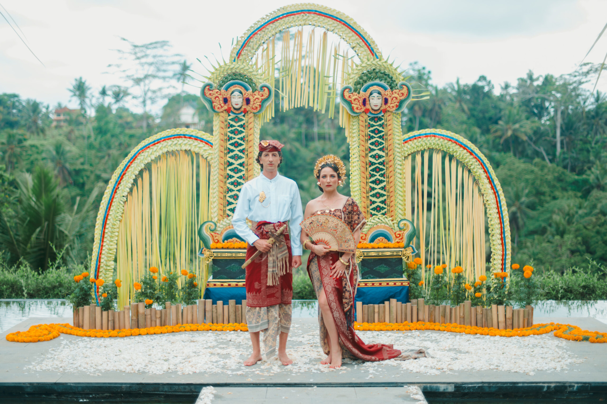 Wedding couple in traditional Balinese attire stands before a colorful ceremonial altar in a lush tropical setting.