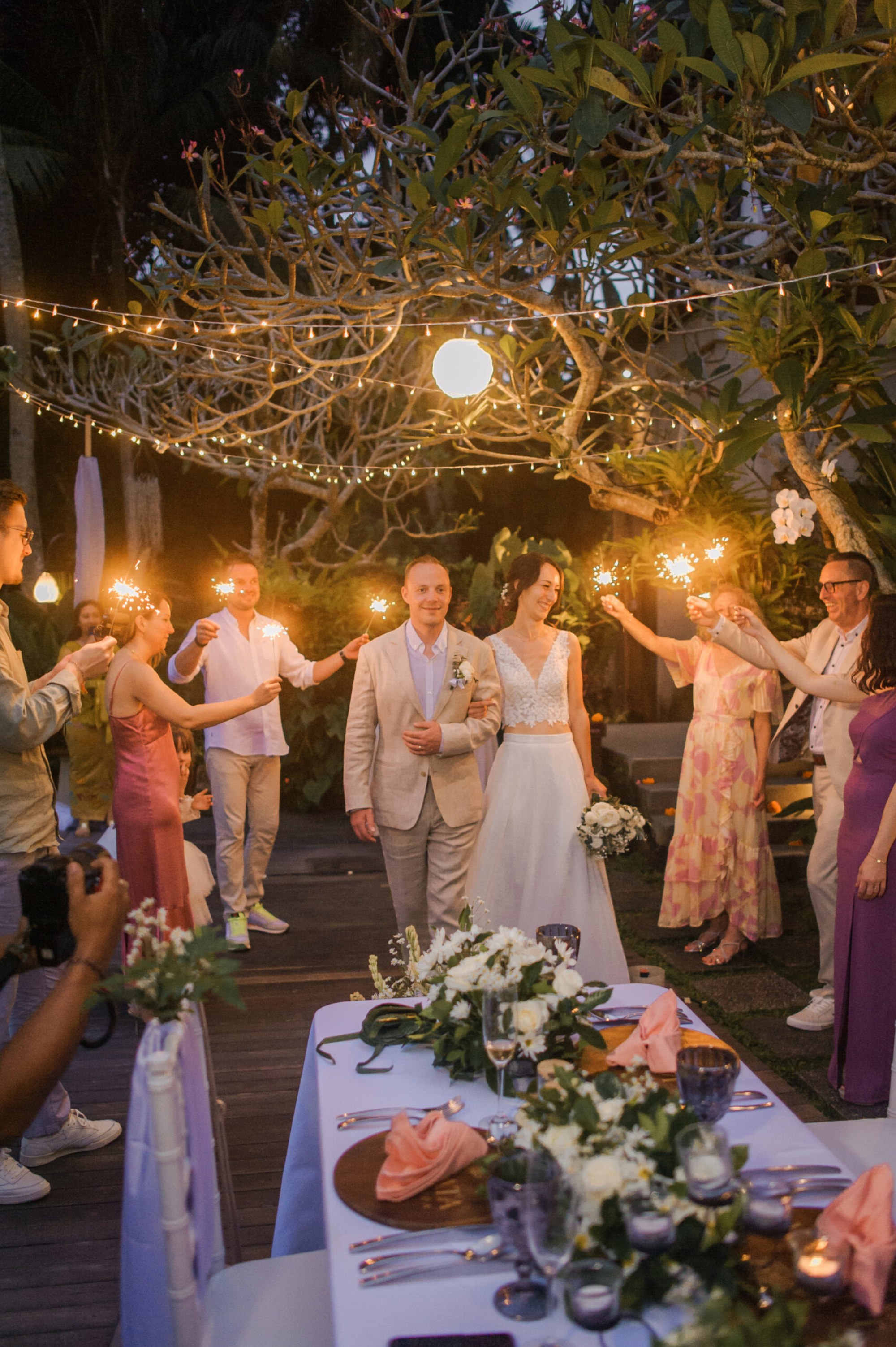 Bride and groom walk through guests holding sparklers at an intimate evening garden wedding reception.