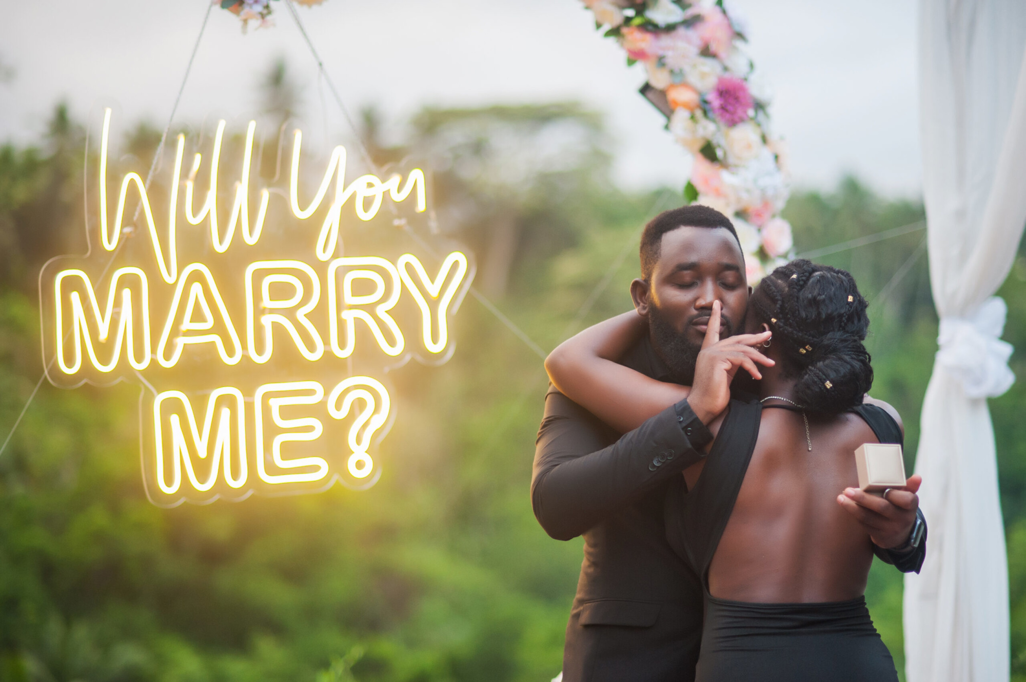 Romantic outdoor proposal with a glowing 'Will You Marry Me?' neon sign and a couple embracing.