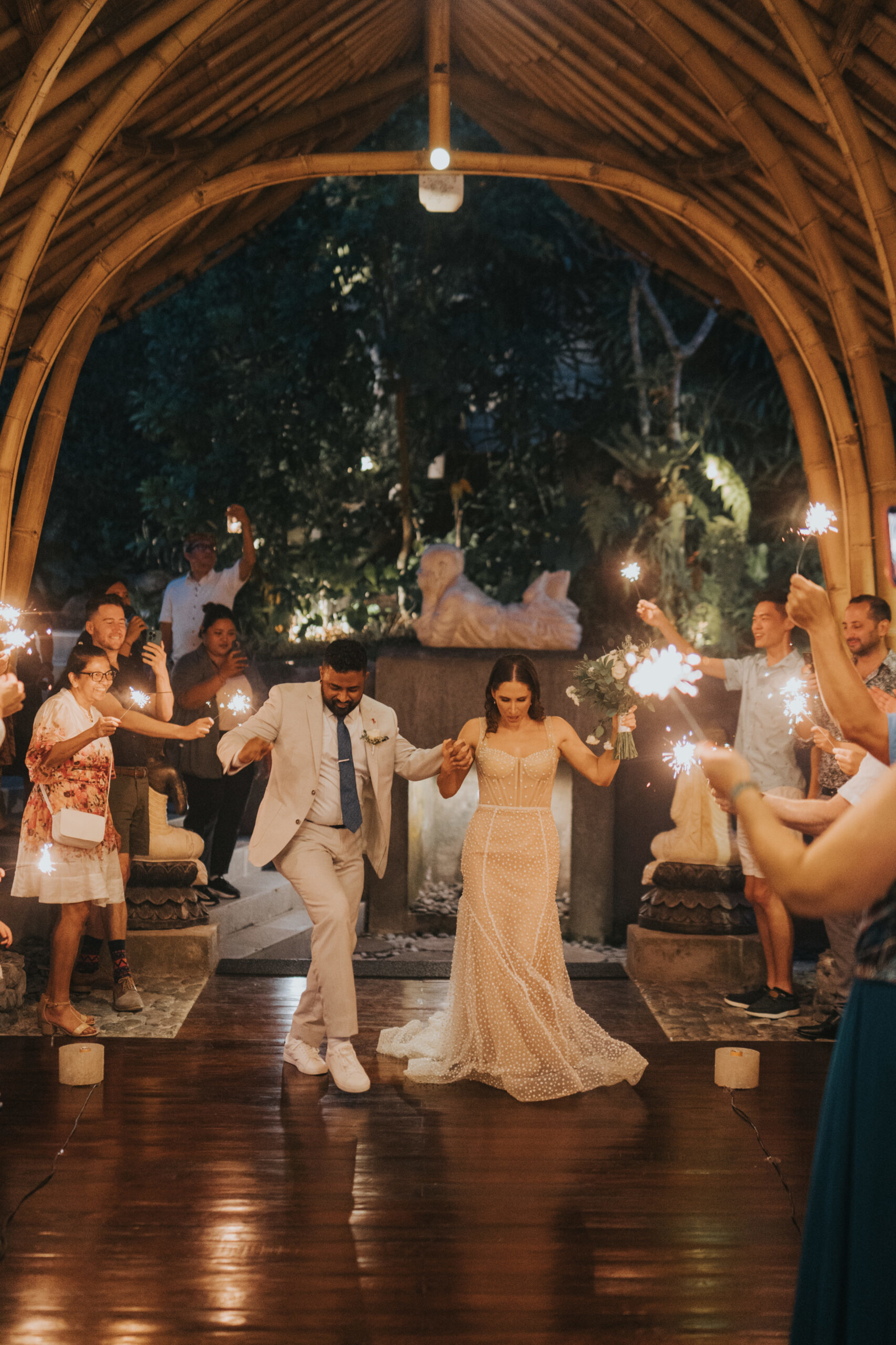 Bride and groom make a joyful sparkler exit under a bamboo pavilion surrounded by cheering guests.