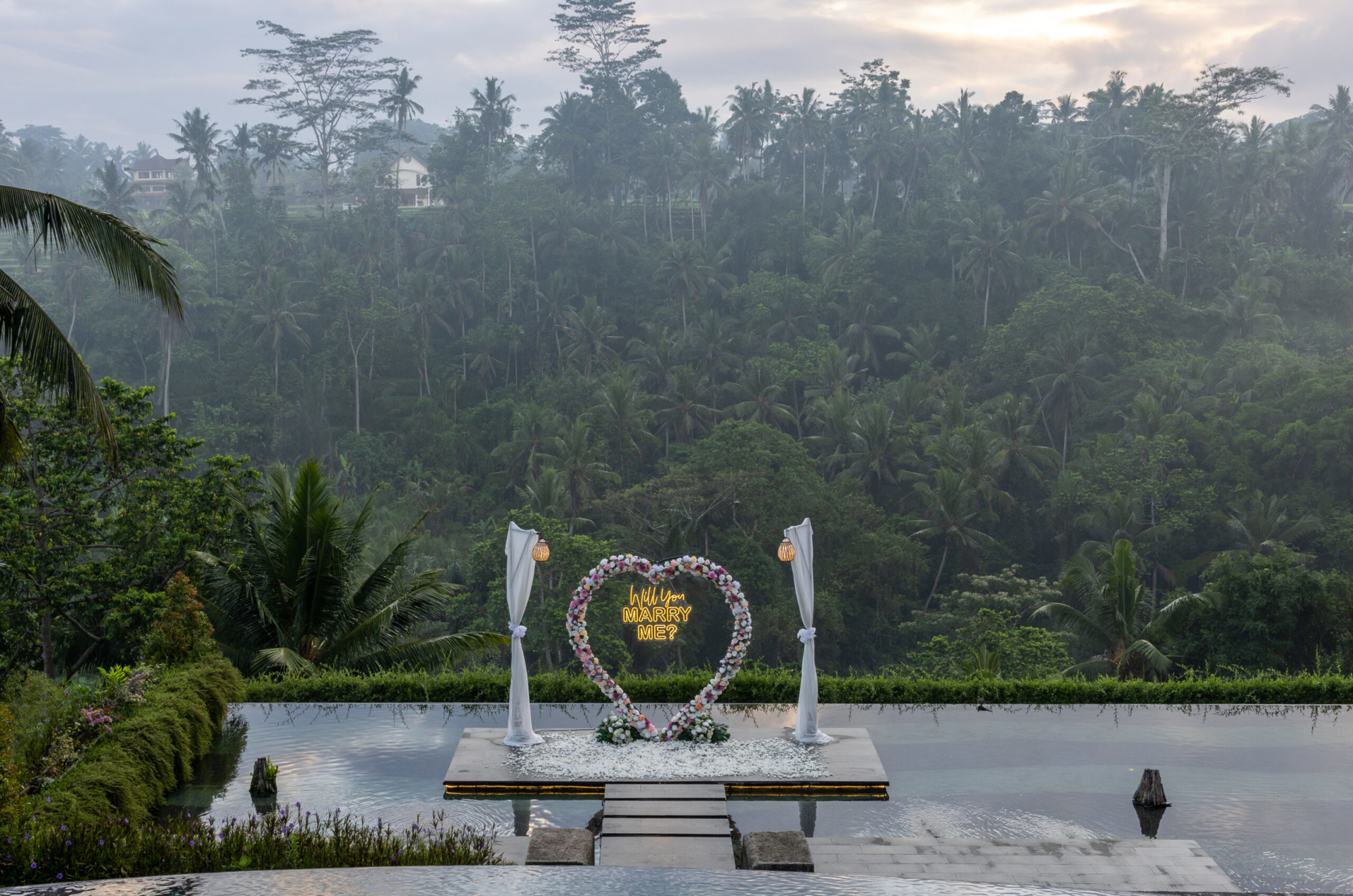 Romantic poolside jungle proposal setup with floral heart arch and neon Will You Marry Me sign at sunset.