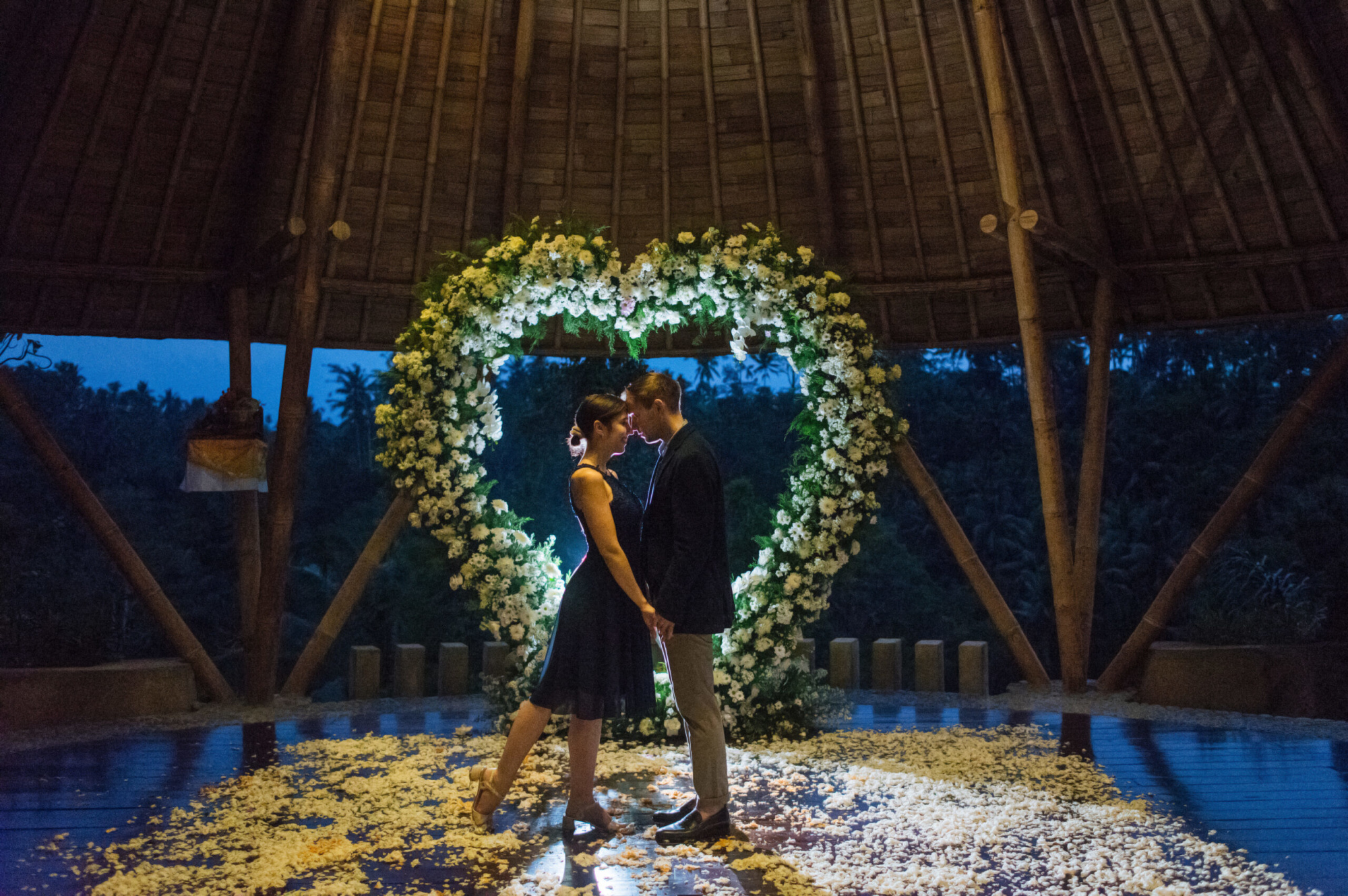 Couple stands embraced under a heart-shaped floral arch at an intimate evening wedding ceremony.