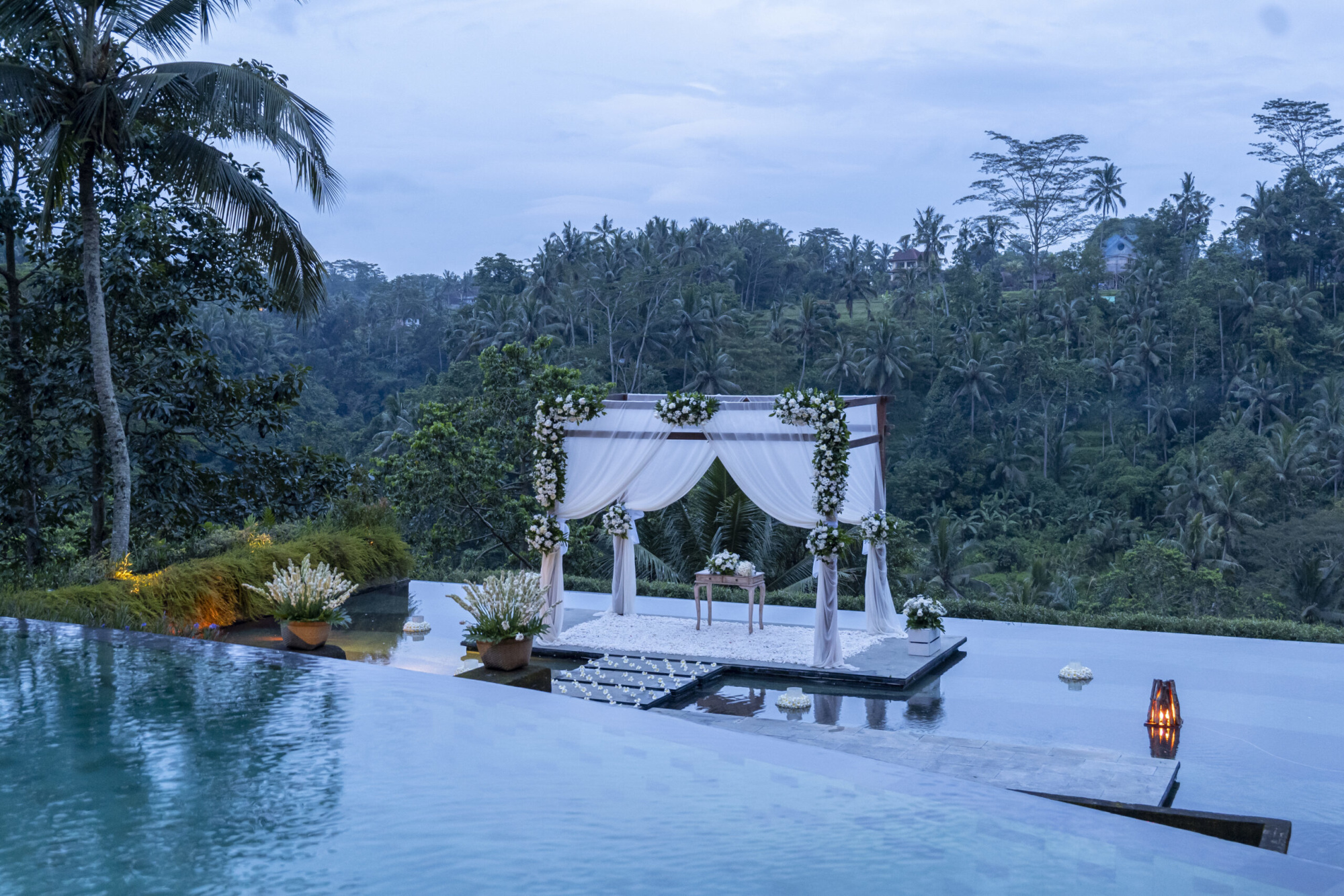 Elegant poolside wedding ceremony arch draped in white fabric overlooking a lush tropical jungle valley.