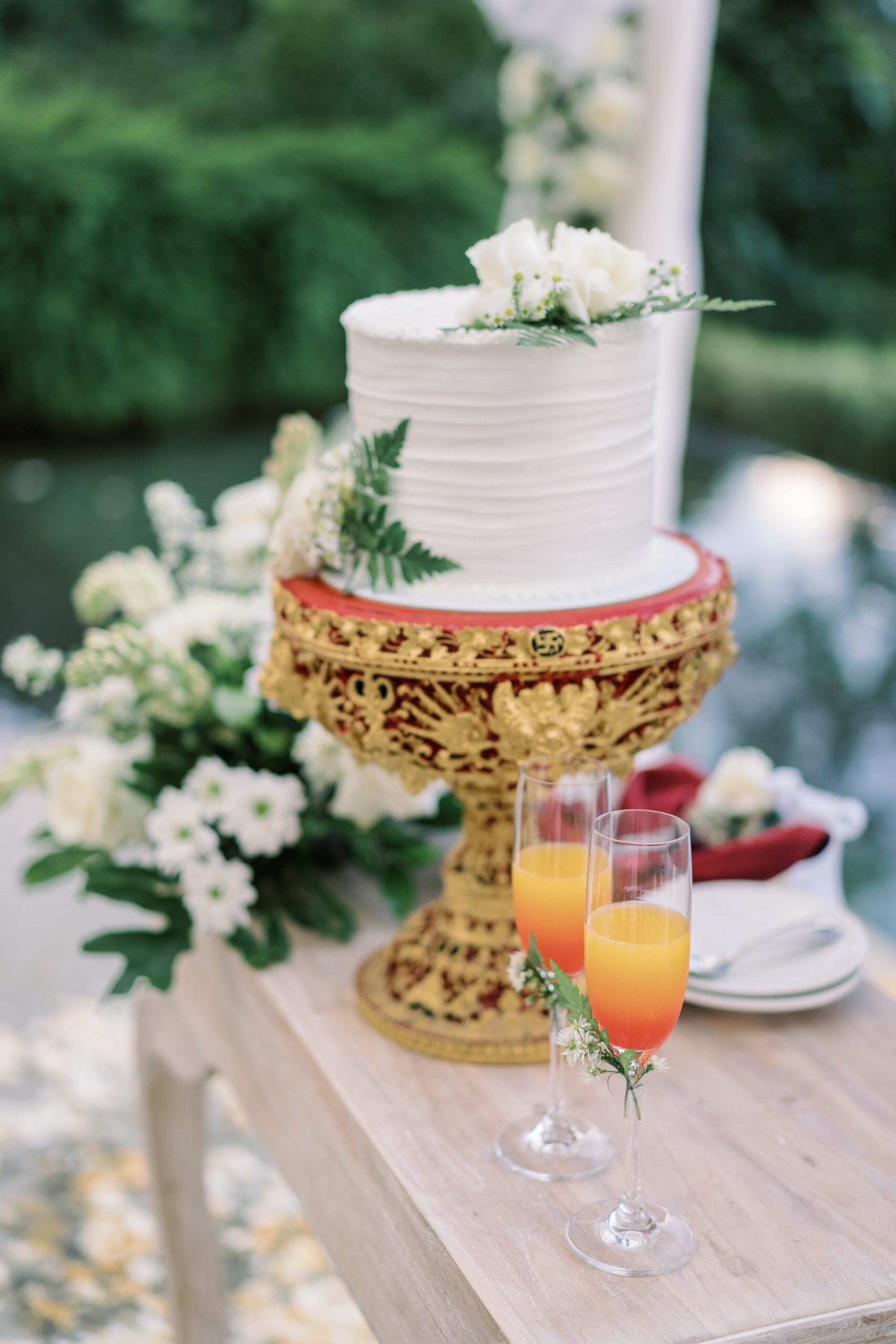 Elegant white wedding cake on ornate gold stand with floral accents and two orange cocktails on a wooden table outdoors.