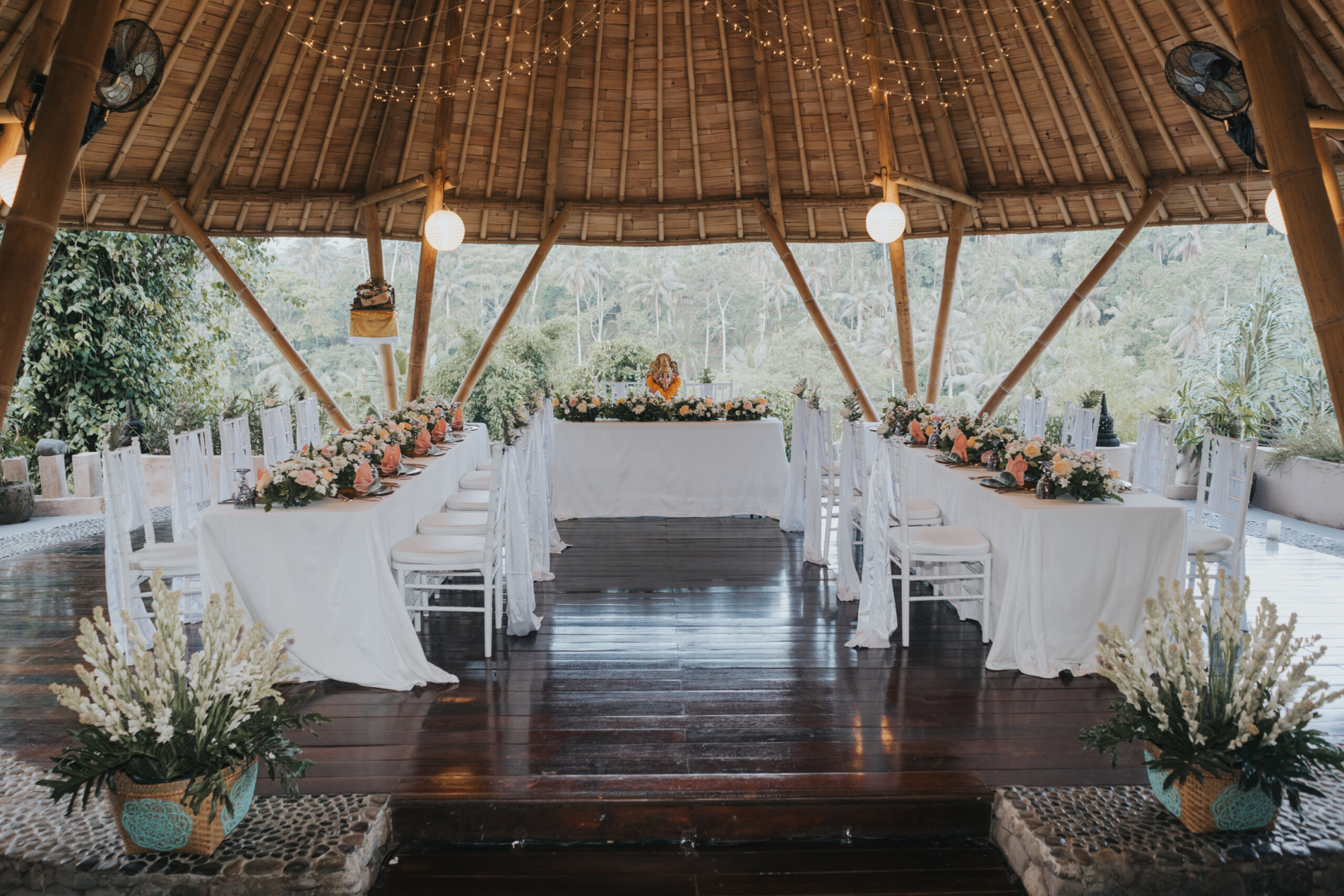 Elegant wedding reception setup under a bamboo pavilion with long floral tables and string lights overlooking lush greenery.