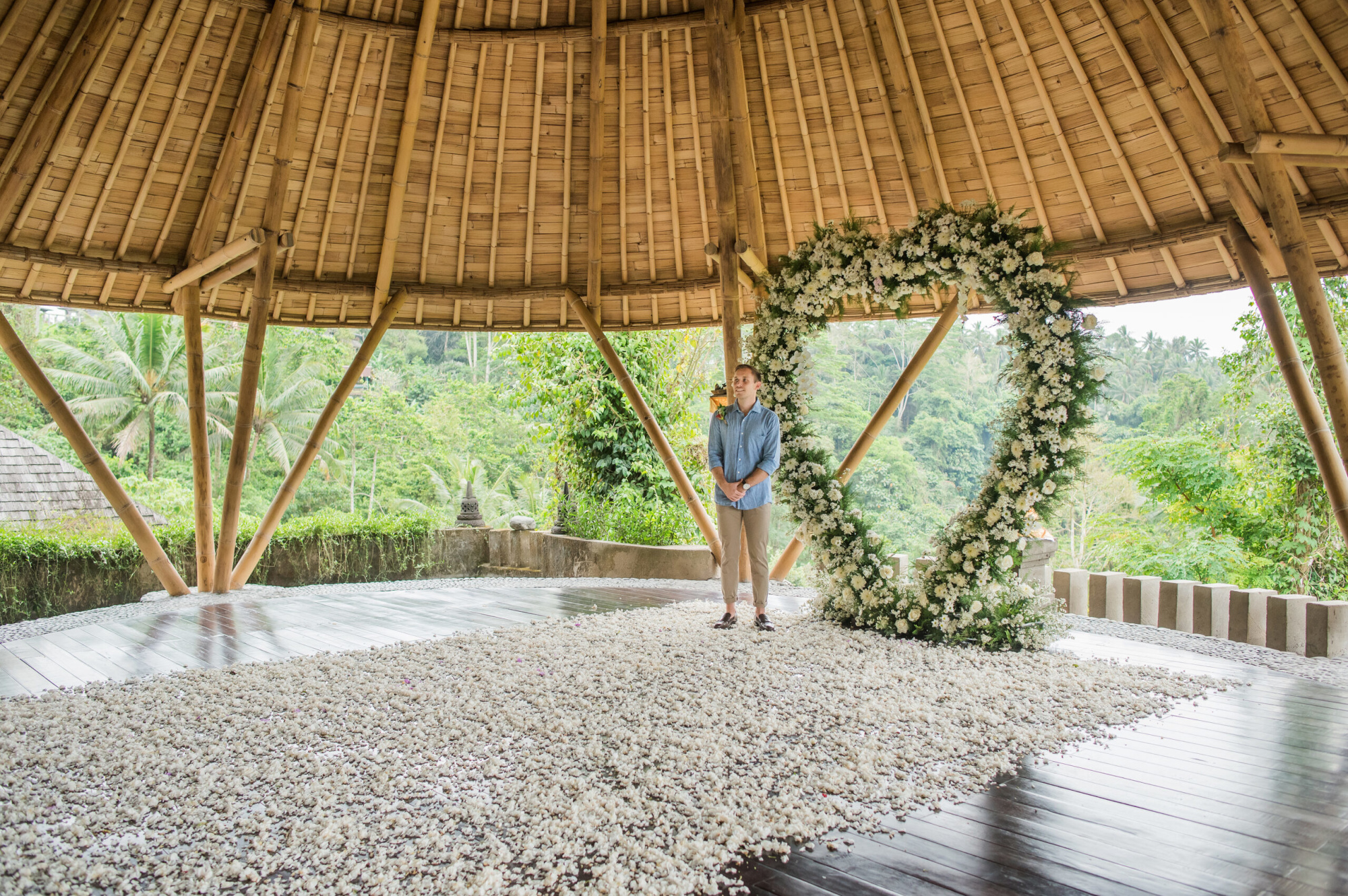 Person standing beneath a large heart-shaped white flower arch in a bamboo pavilion overlooking lush tropical greenery.