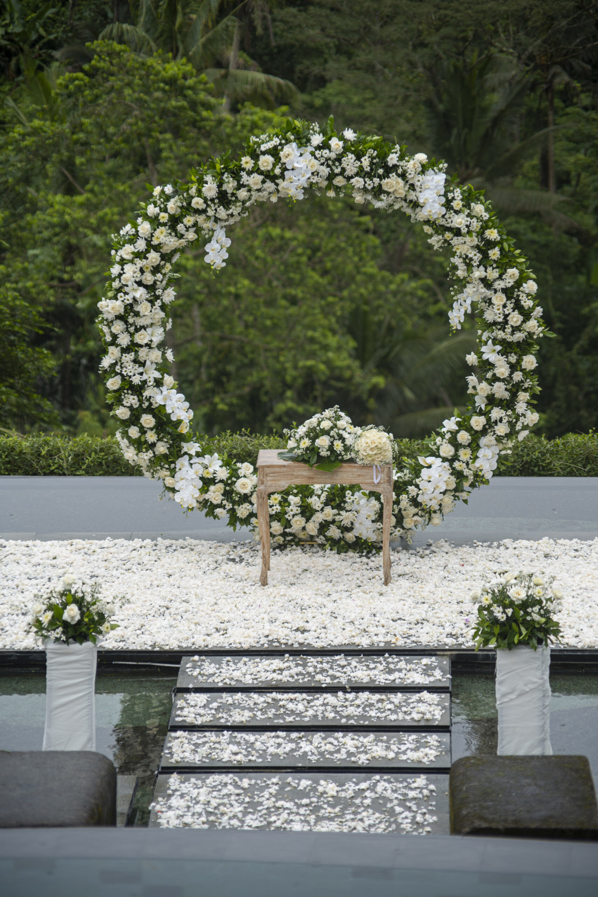 Outdoor wedding ceremony setup with a large circular white floral arch and petals lining the aisle over water.