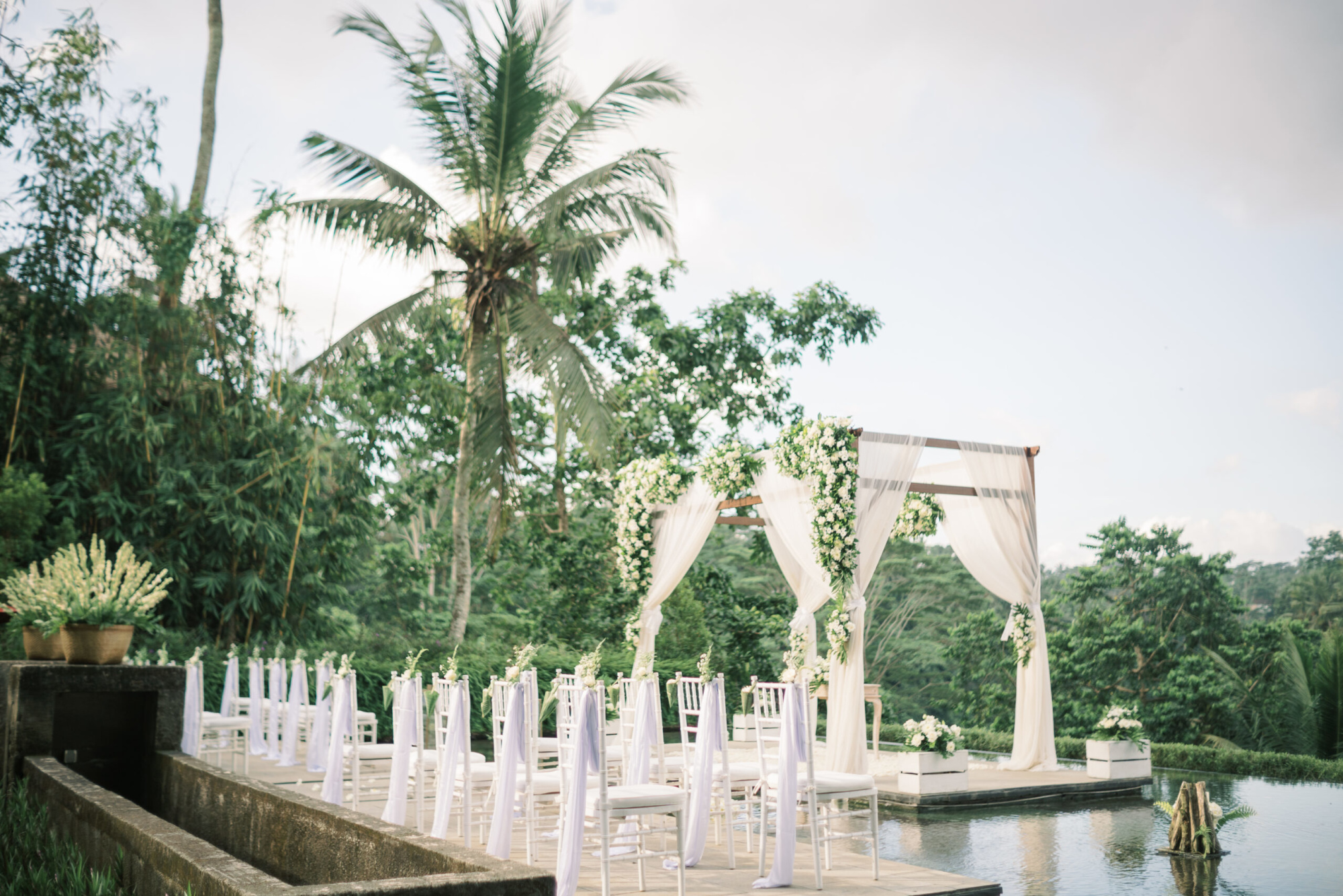 Outdoor tropical wedding ceremony setup with a floral draped arch and white chairs beside a reflective pool.