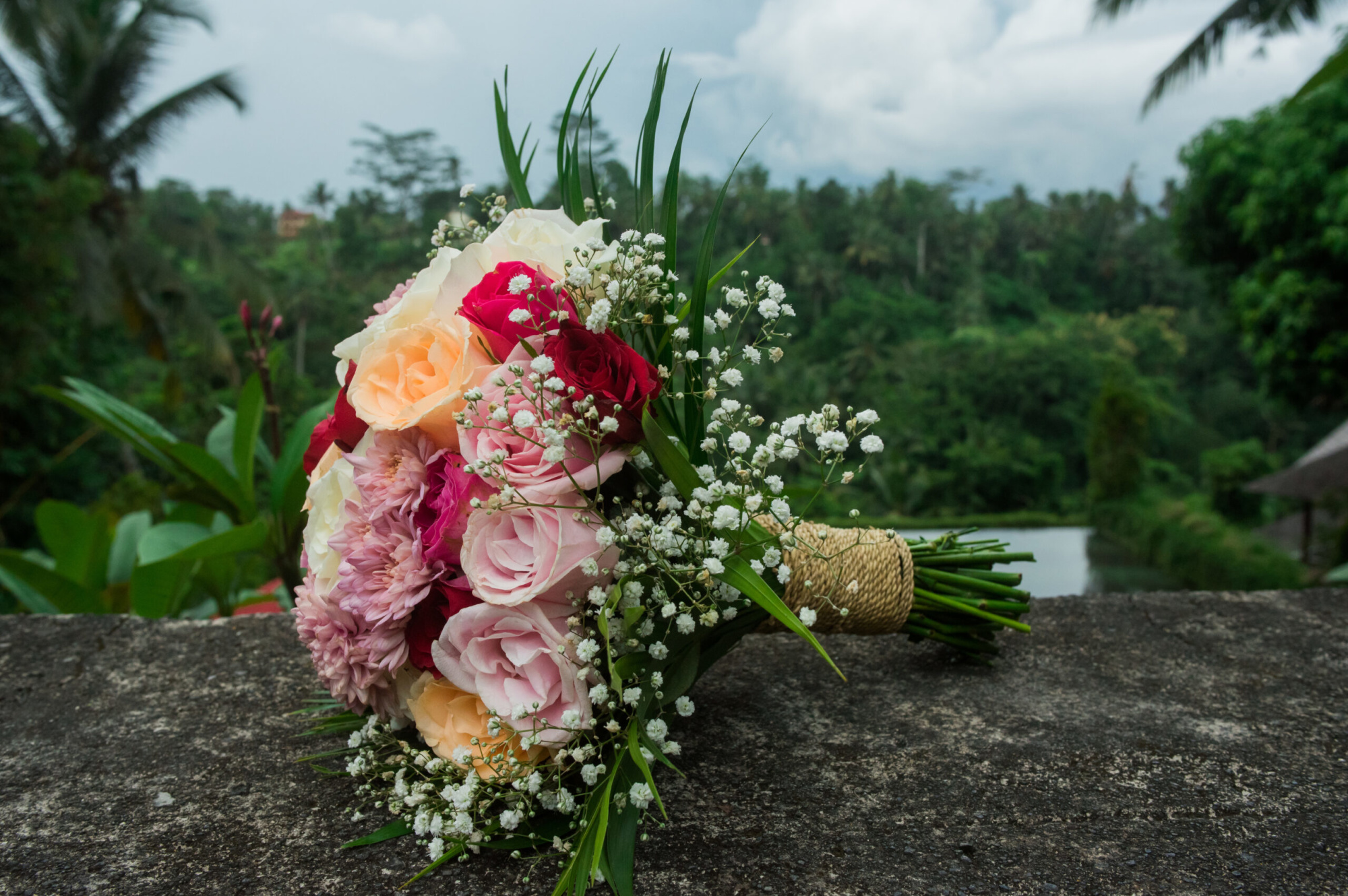 Romantic bridal bouquet of pink, peach, and red flowers resting on a stone ledge with lush greenery in the background.