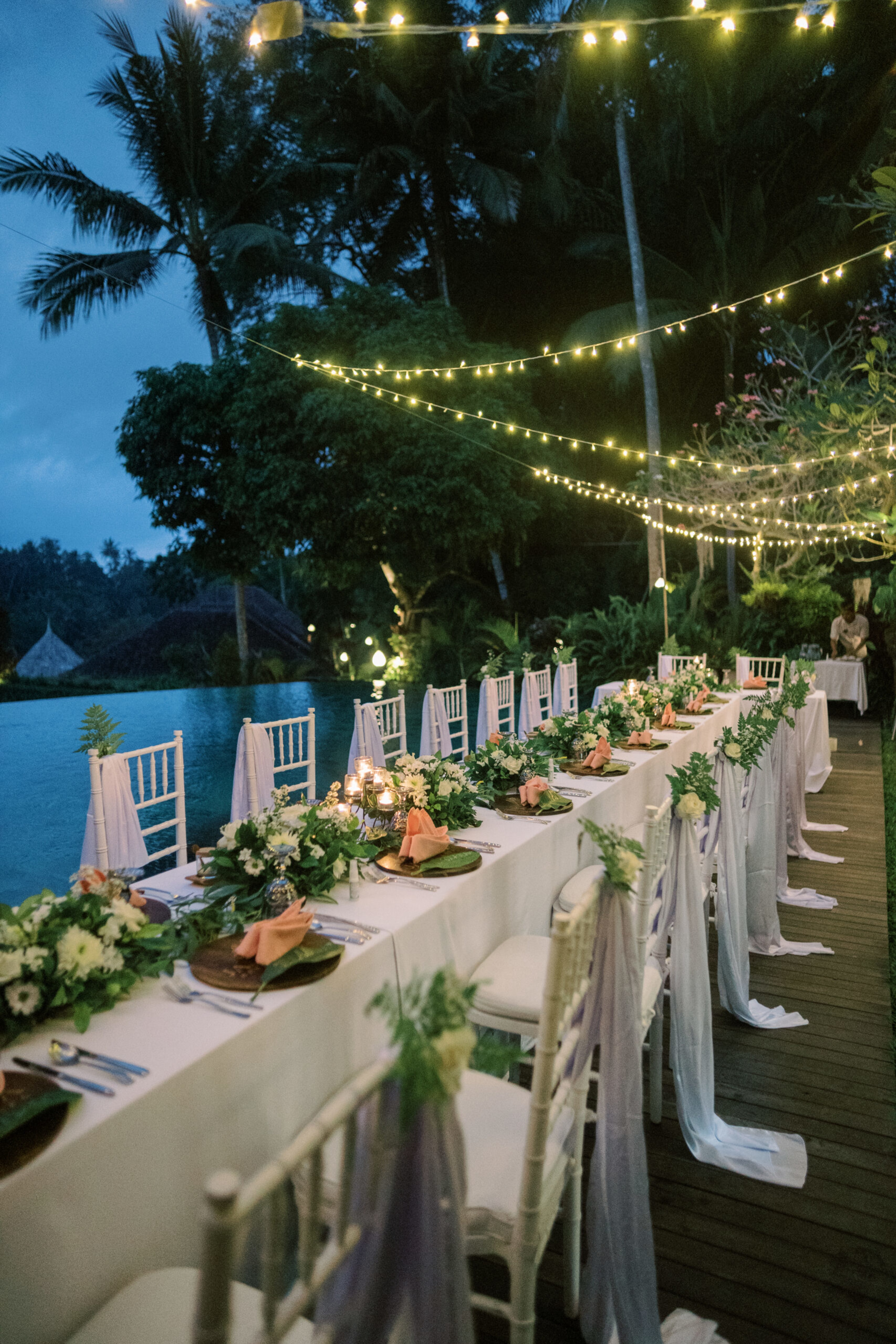 Evening outdoor wedding reception with long decorated tables by a pool under glowing string lights.