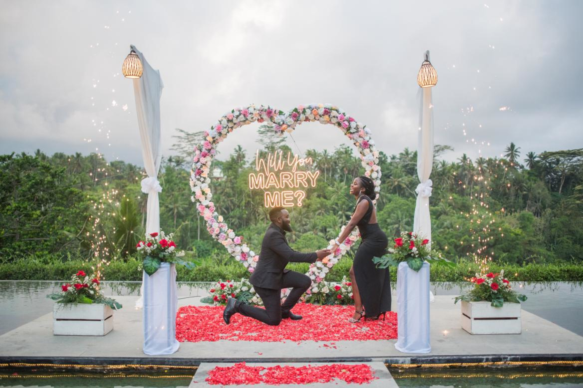 Romantic outdoor proposal with man kneeling under floral heart arch and neon Will You Marry Me sign surrounded by rose petals and sparklers.