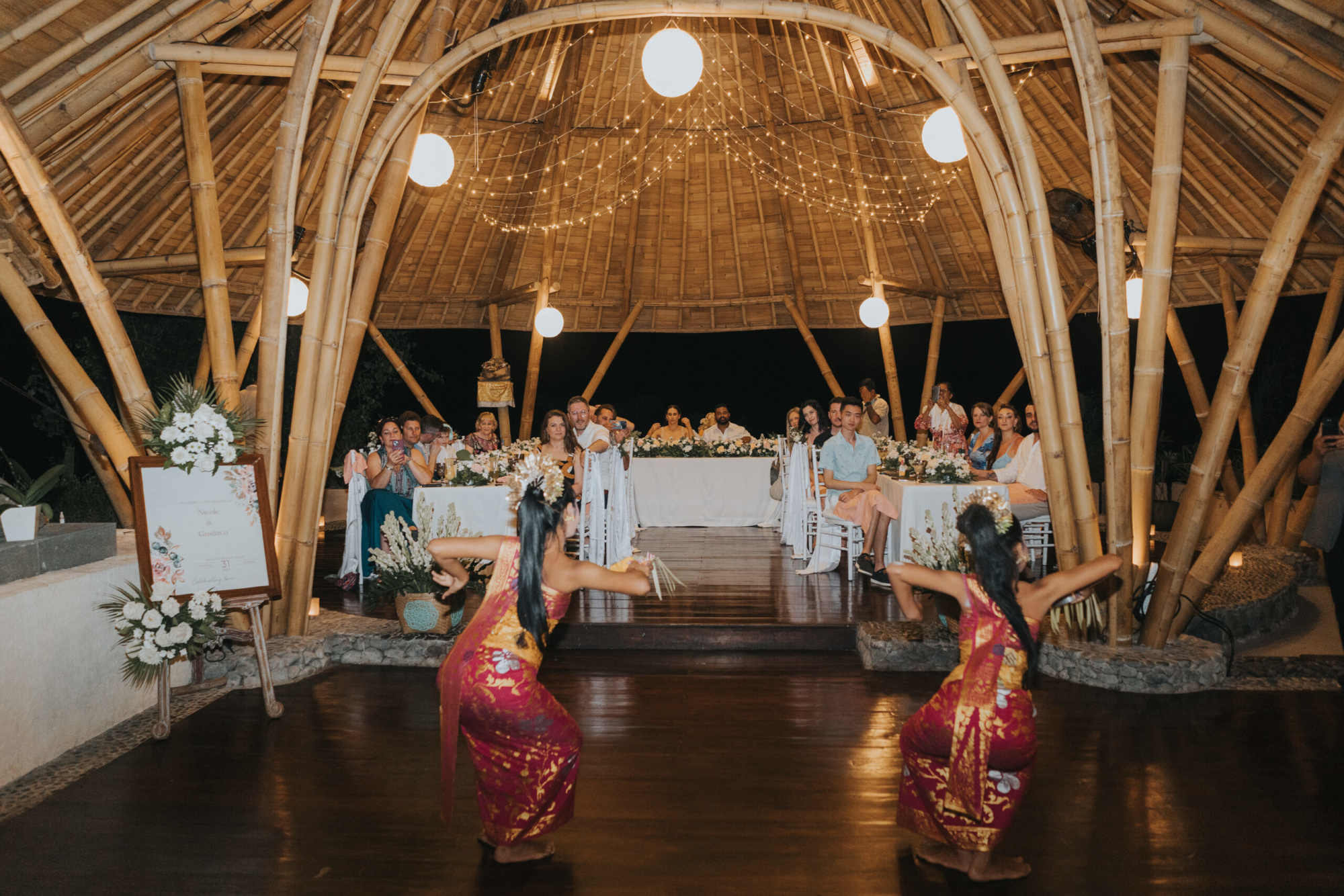 Wedding guests watch traditional dancers perform in a bamboo pavilion decorated with flowers and string lights.