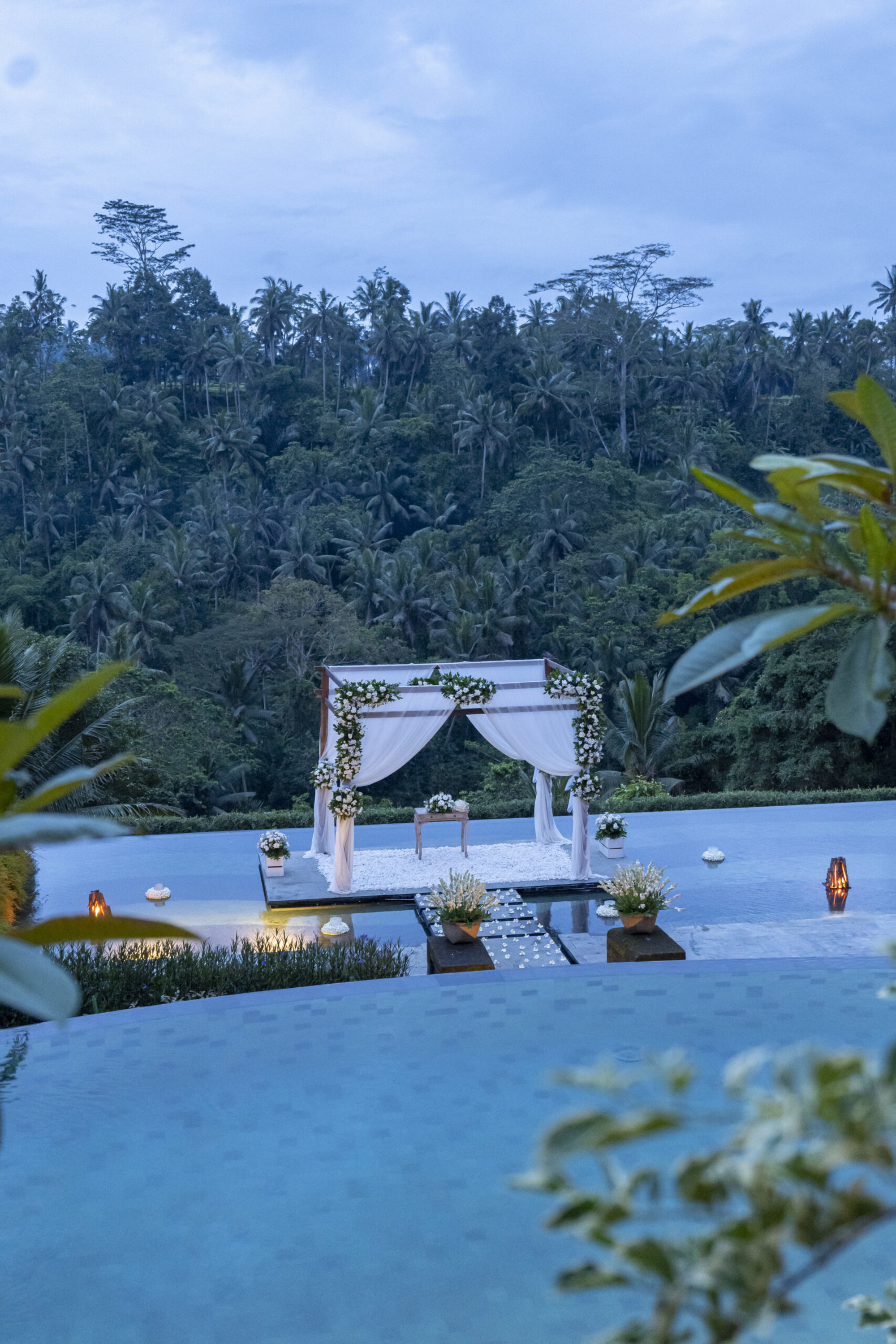 Tropical poolside wedding ceremony arch draped in white fabric and flowers overlooking lush green jungle hills.
