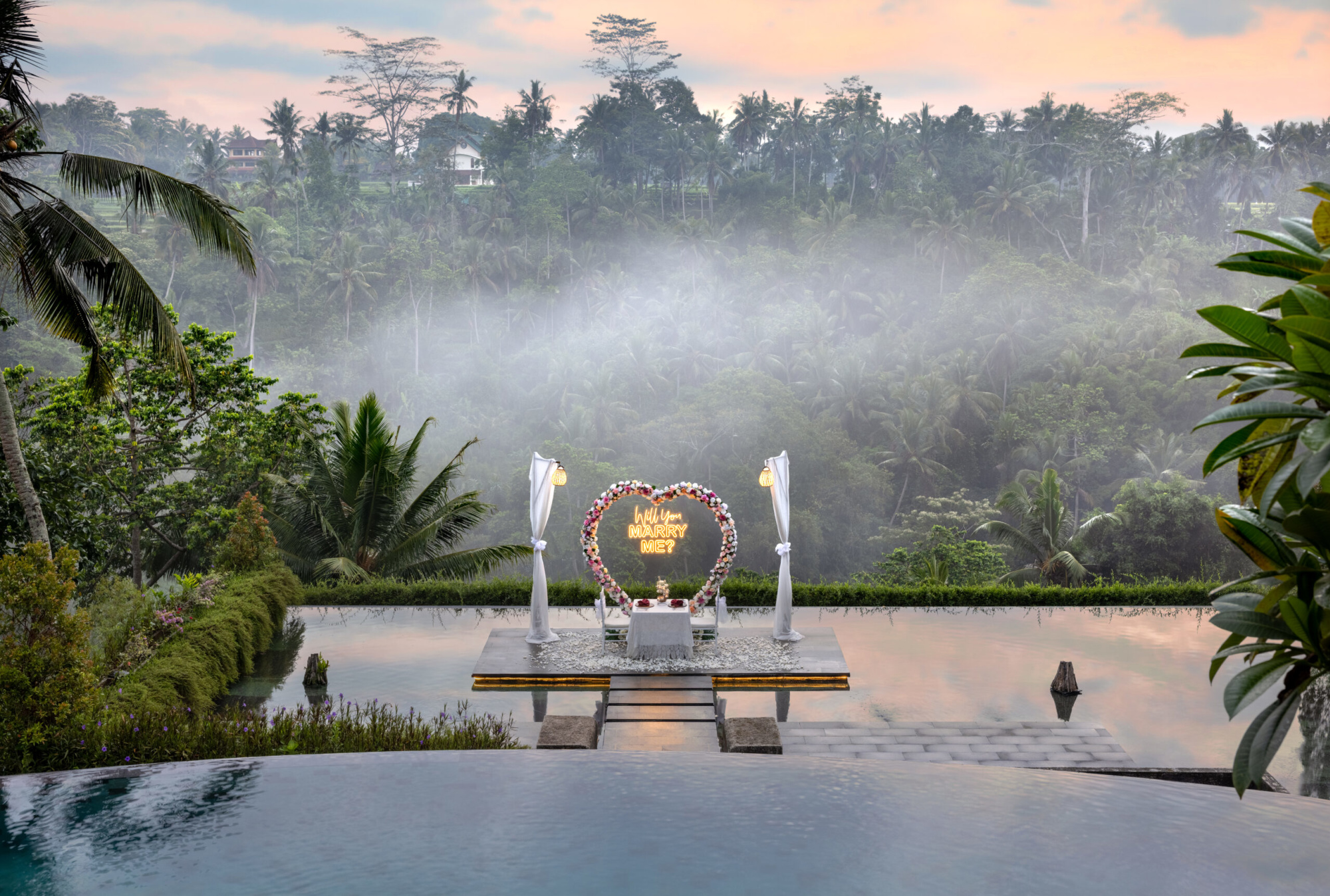 Romantic proposal setup by an infinity pool overlooking a misty tropical jungle at sunset.