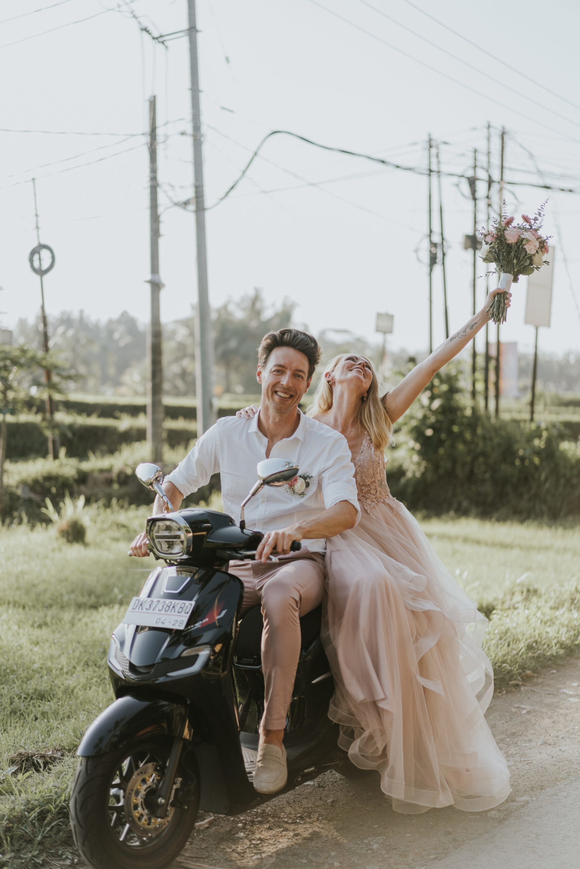Joyful wedding couple rides a scooter down a sunlit country road, the bride lifting her bouquet in celebration.