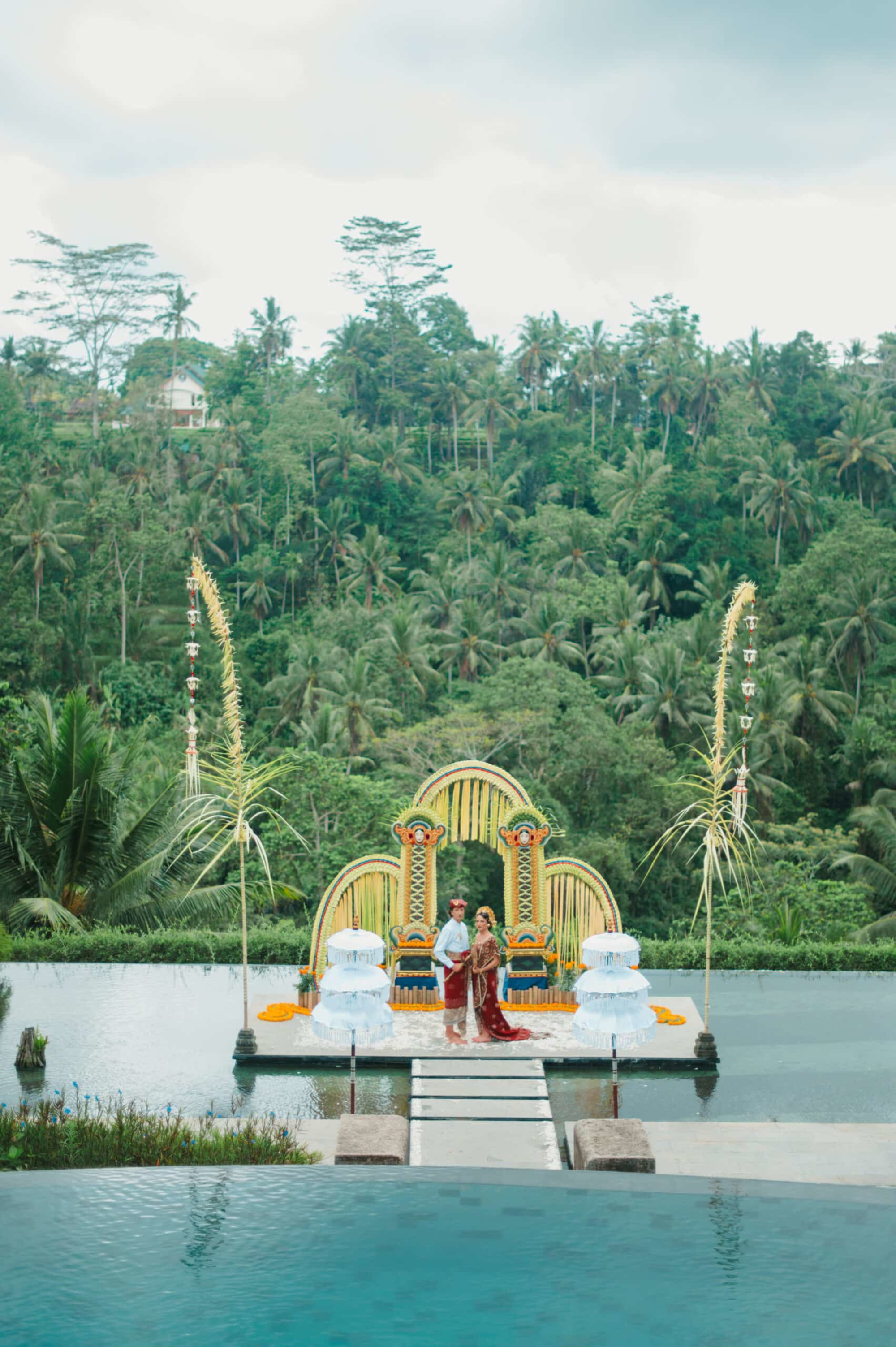 Couple stands on a colorful Balinese wedding altar by an infinity pool with lush jungle hills in the background.