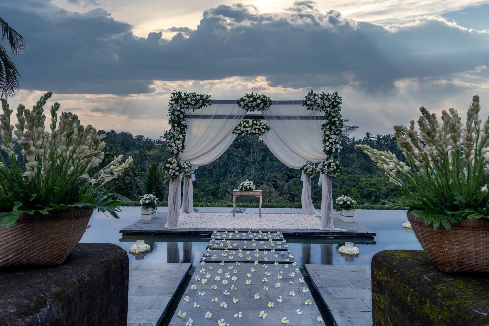 Outdoor wedding ceremony arch with white drapery and flowers overlooking a lush tropical landscape at sunset.