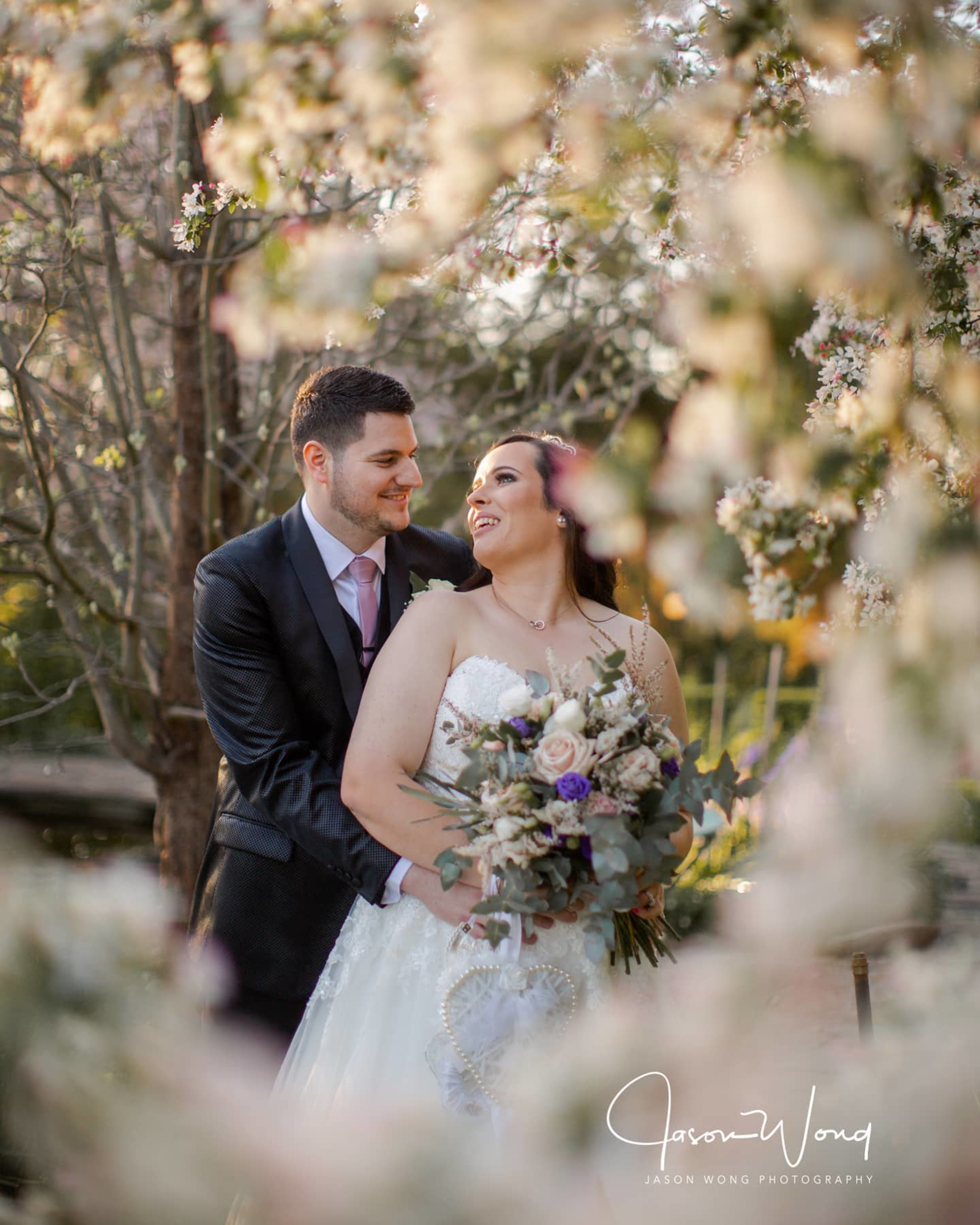 Bride and groom embrace in a blooming garden framed by soft spring blossoms at golden hour.