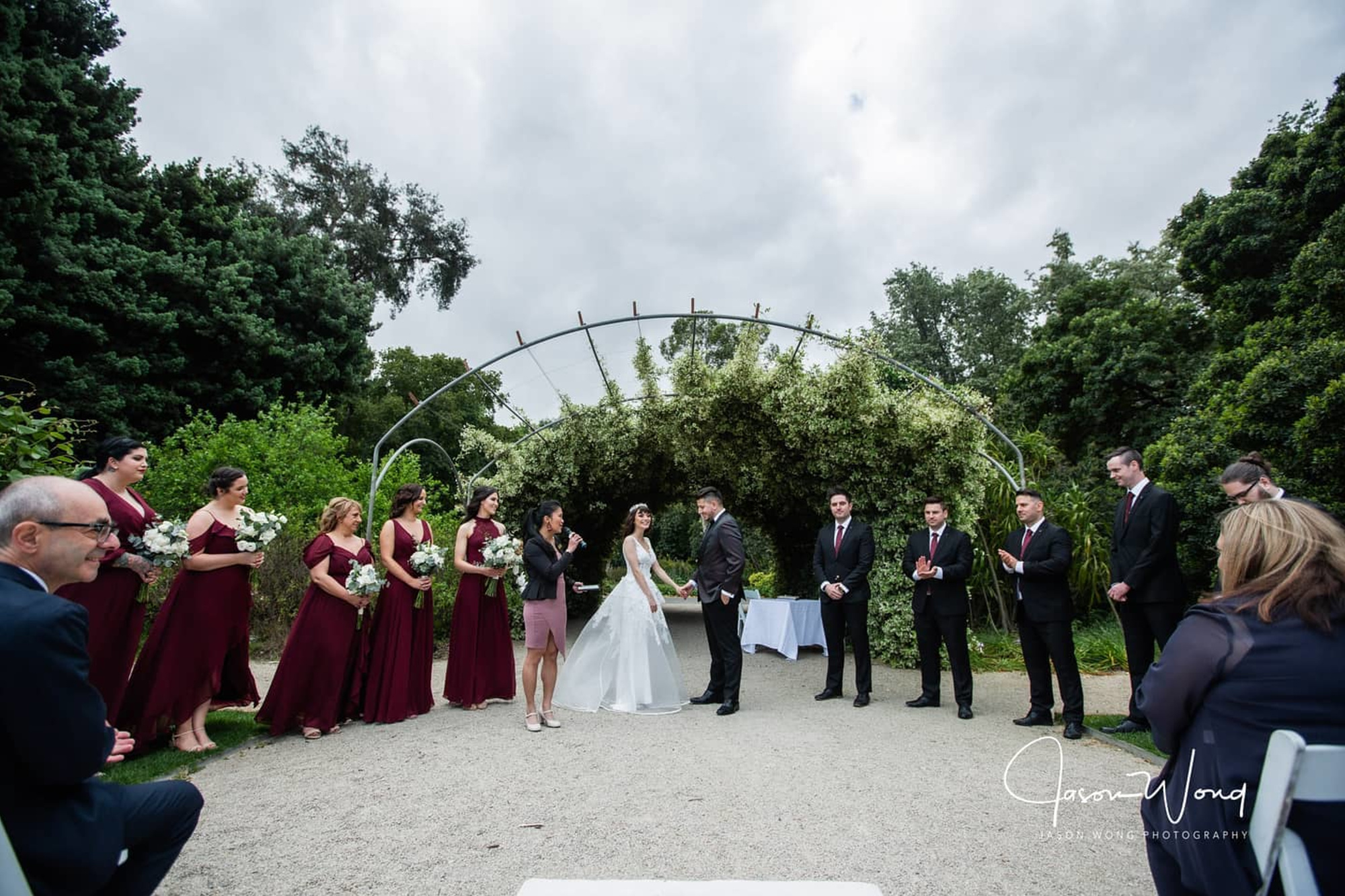 Outdoor garden wedding ceremony with bride, groom, and bridal party standing under a lush green archway.