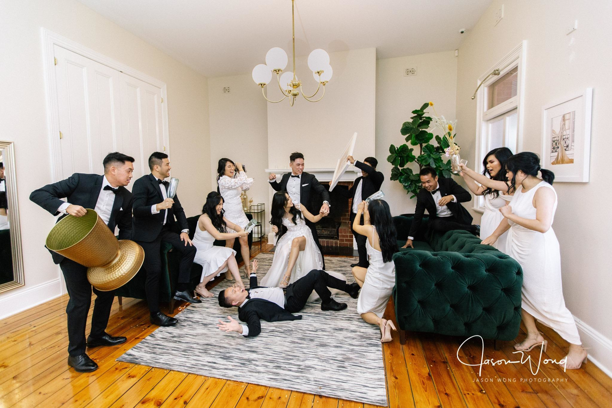 Playful wedding party posing in a bright living room with the couple surrounded by laughing bridesmaids and groomsmen.