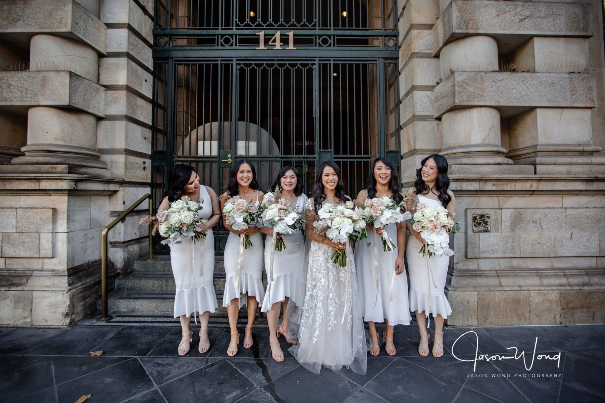 Bride and bridesmaids in white dresses holding pastel bouquets standing and laughing in front of a grand stone building.
