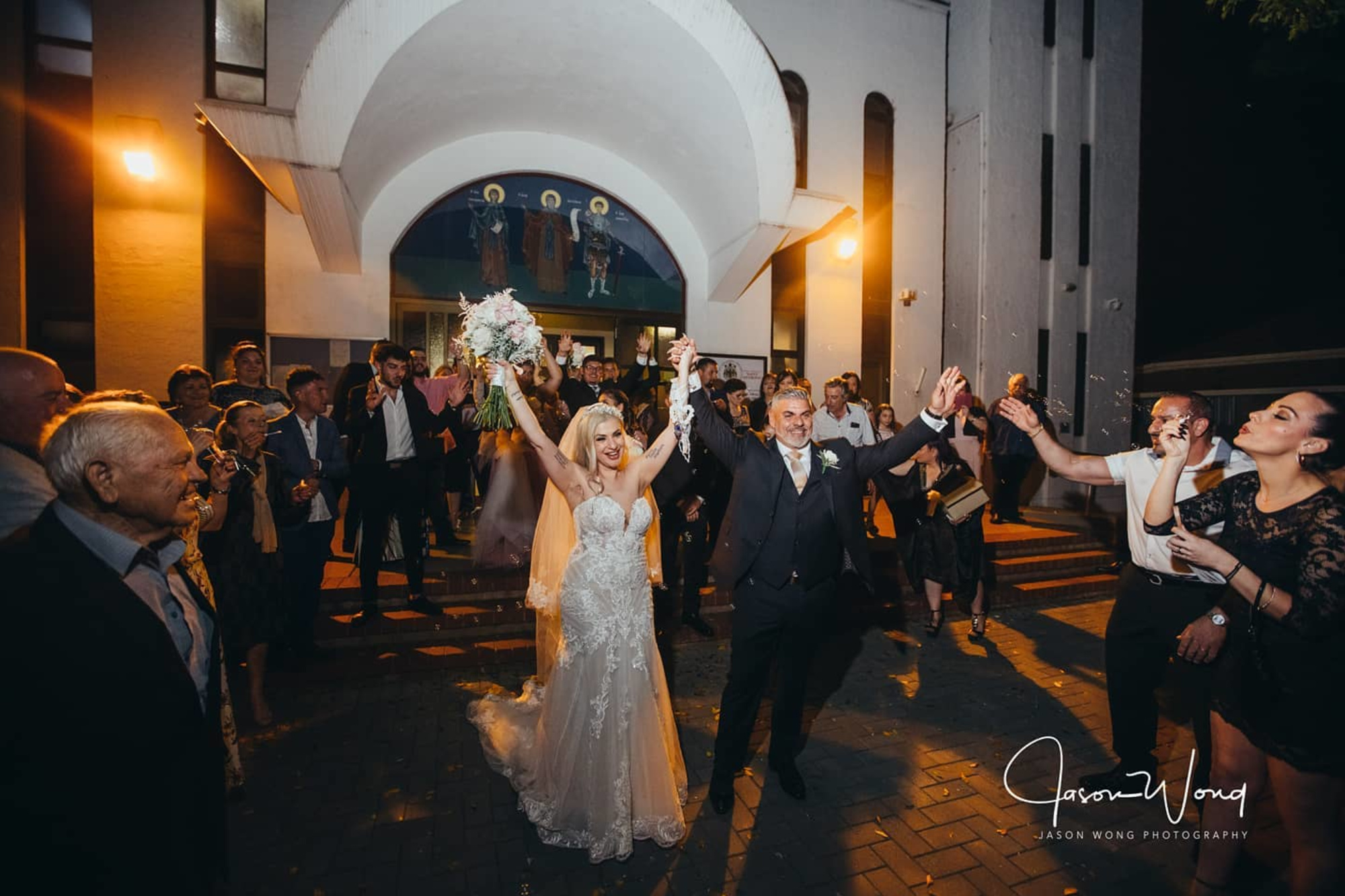 Bride and groom celebrate outside a church at night surrounded by cheering guests.