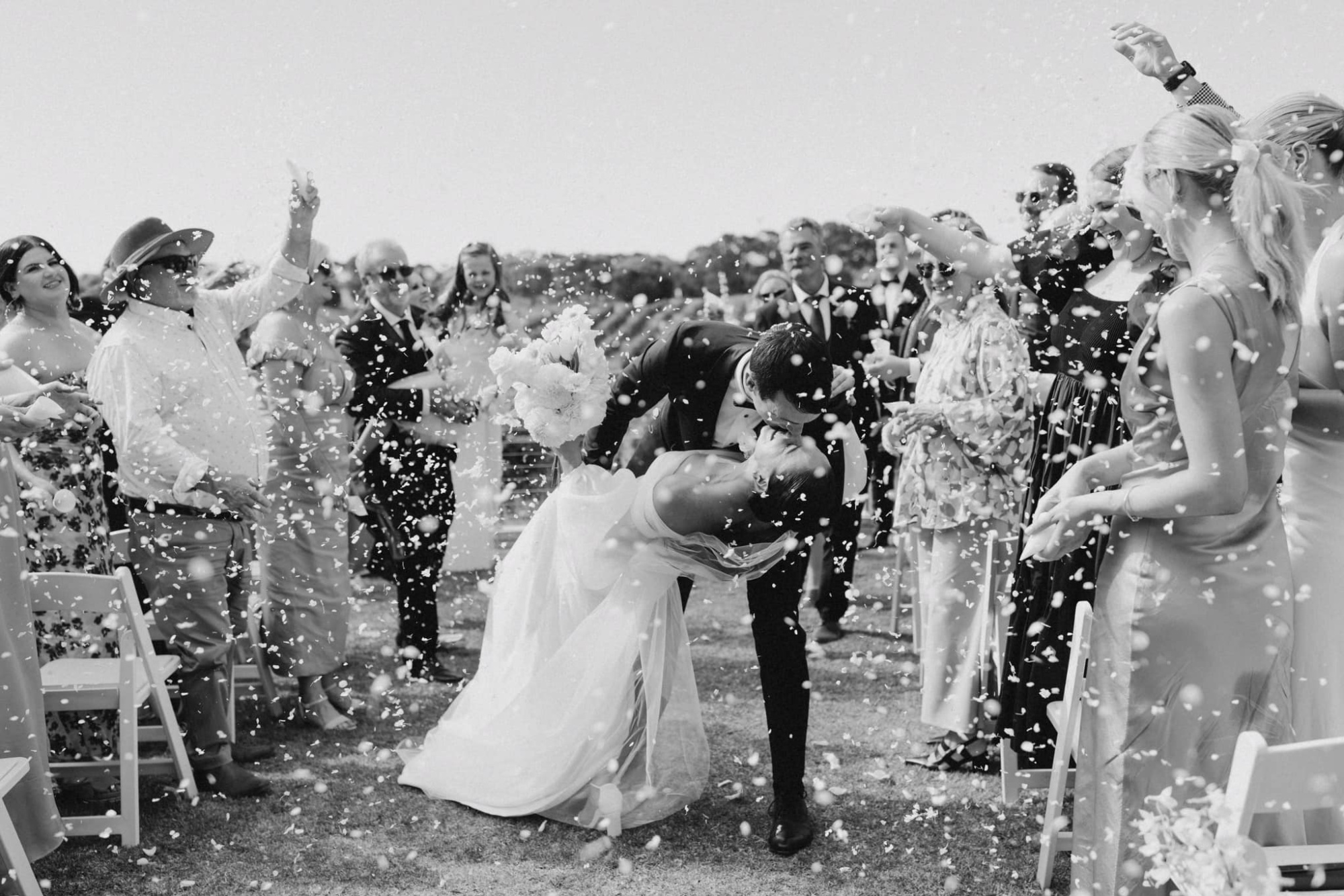 Newlywed couple kisses in a dip while guests shower them with confetti during an outdoor ceremony exit.