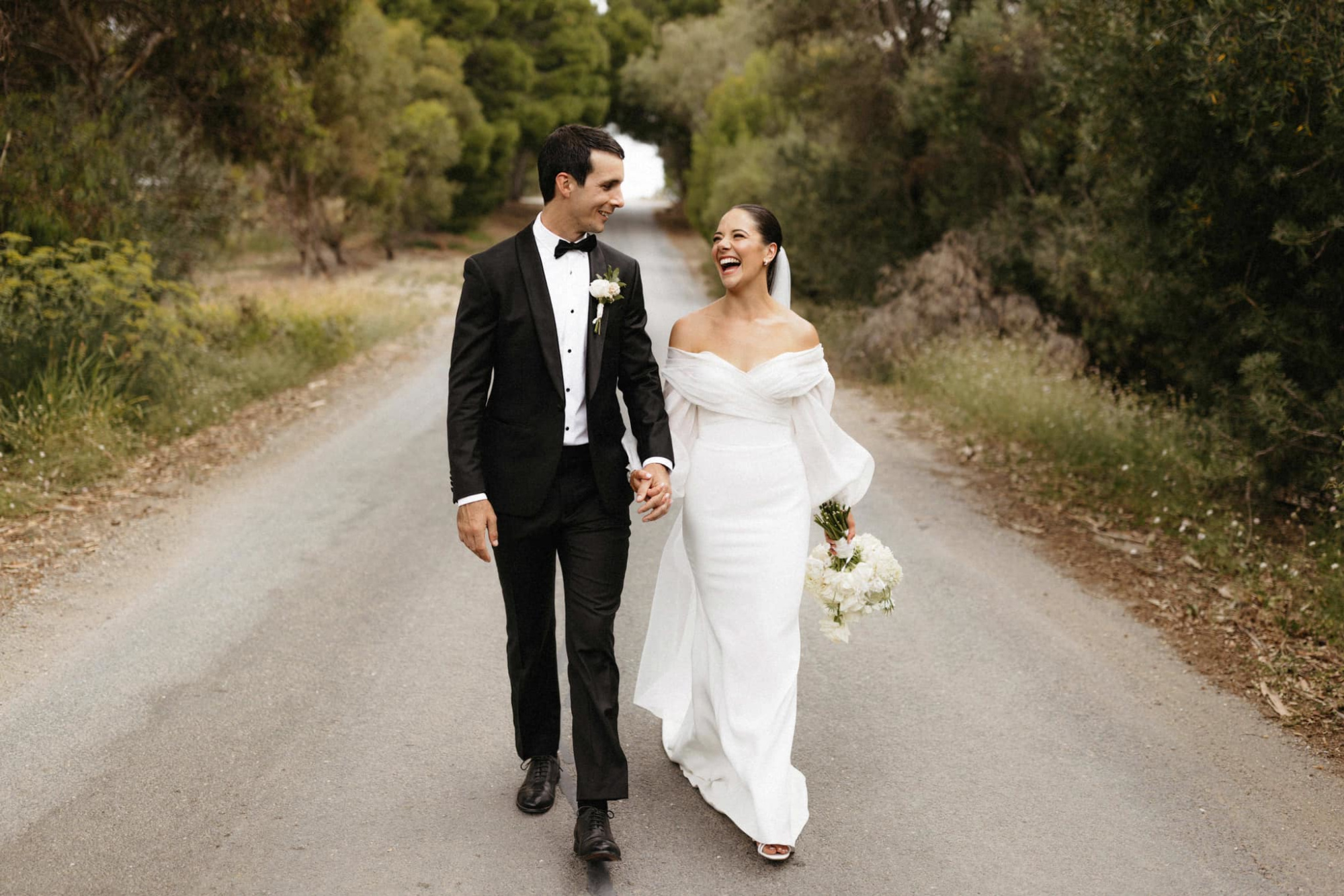 Bride and groom holding hands and smiling while walking down a tree-lined country road.