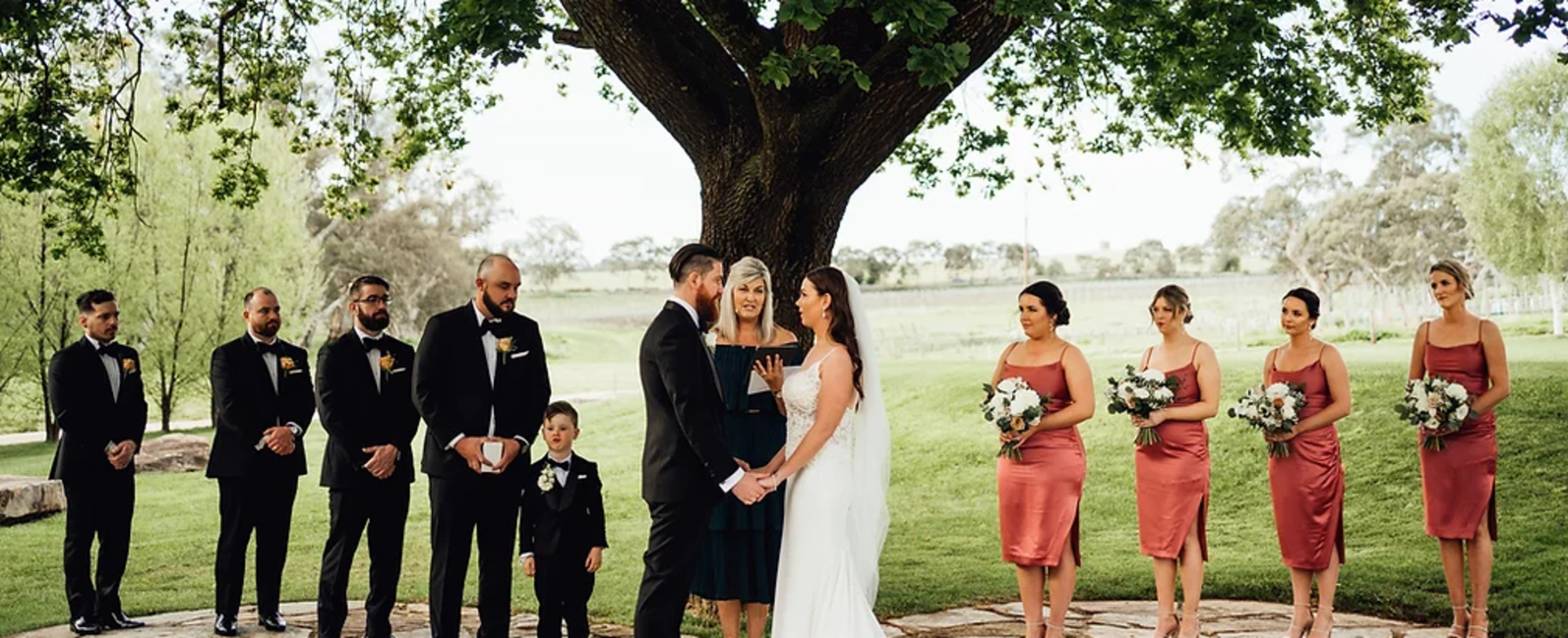 Wedding couple exchange vows under a large tree surrounded by bridesmaids and groomsmen in an outdoor garden setting.