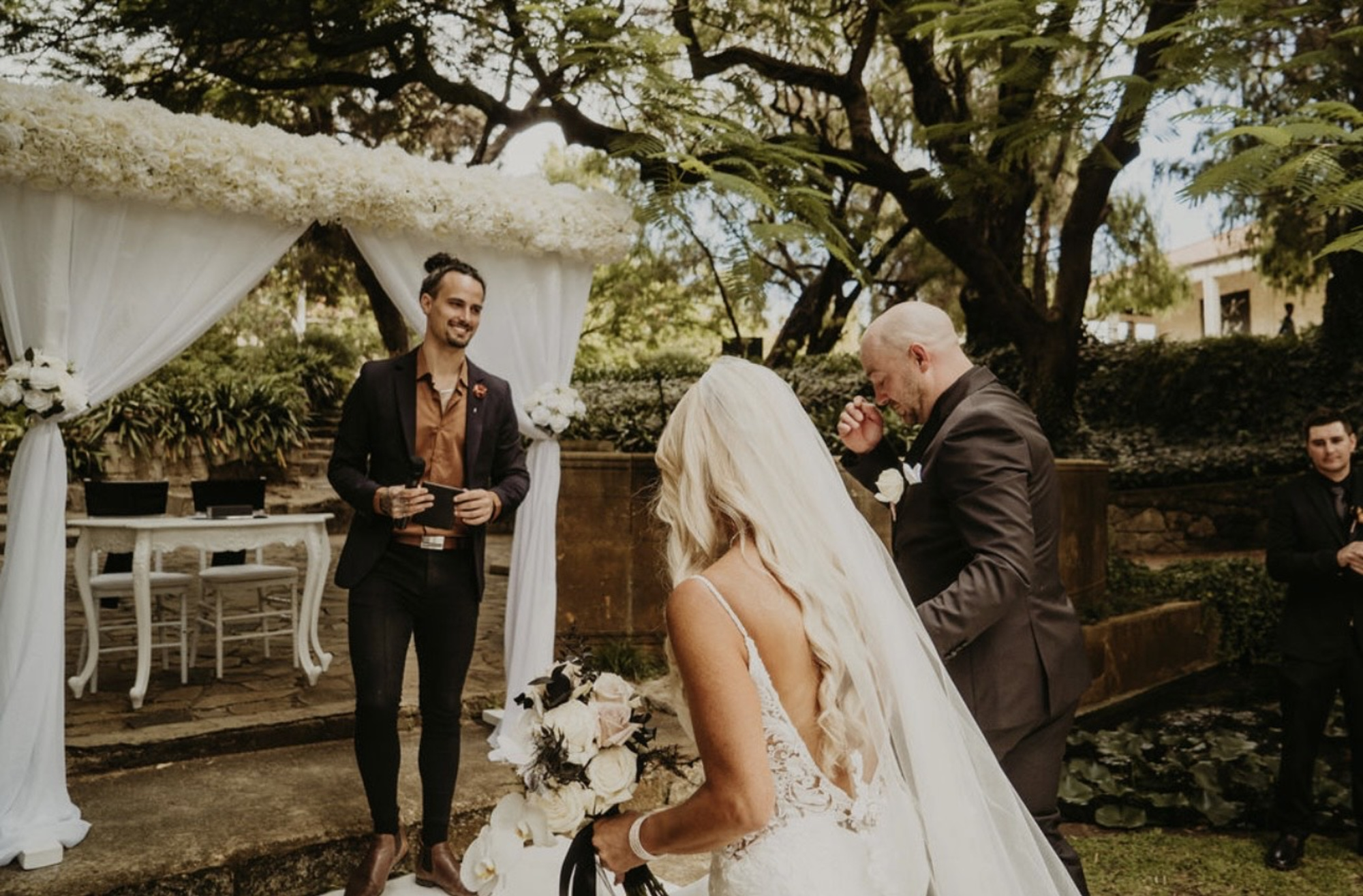 Bride and groom stand before an officiant under a floral-draped arbor in a lush garden ceremony.