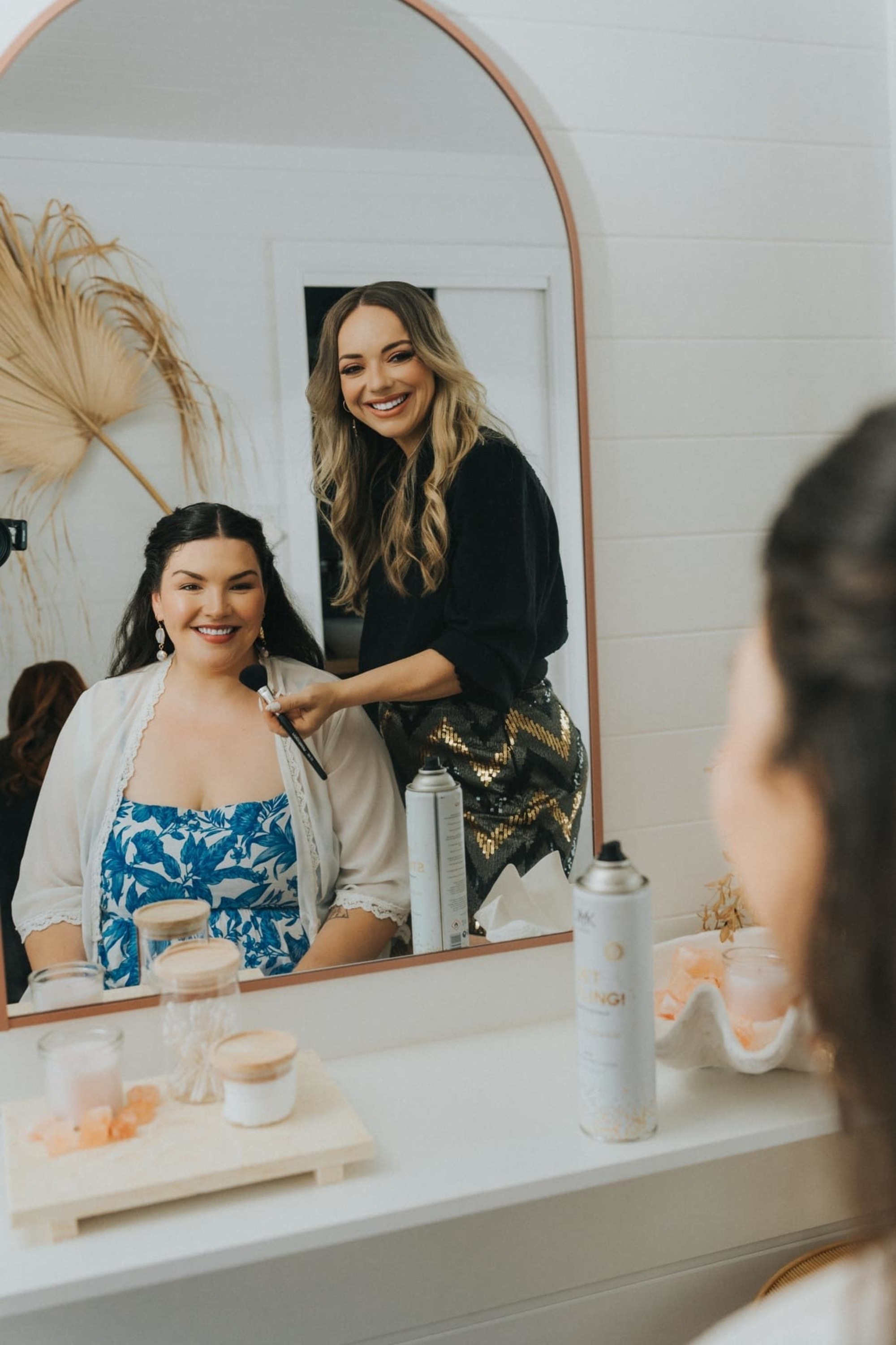 Makeup artist applies bridal makeup as the smiling bride sits in front of a mirror in a bright getting-ready room.
