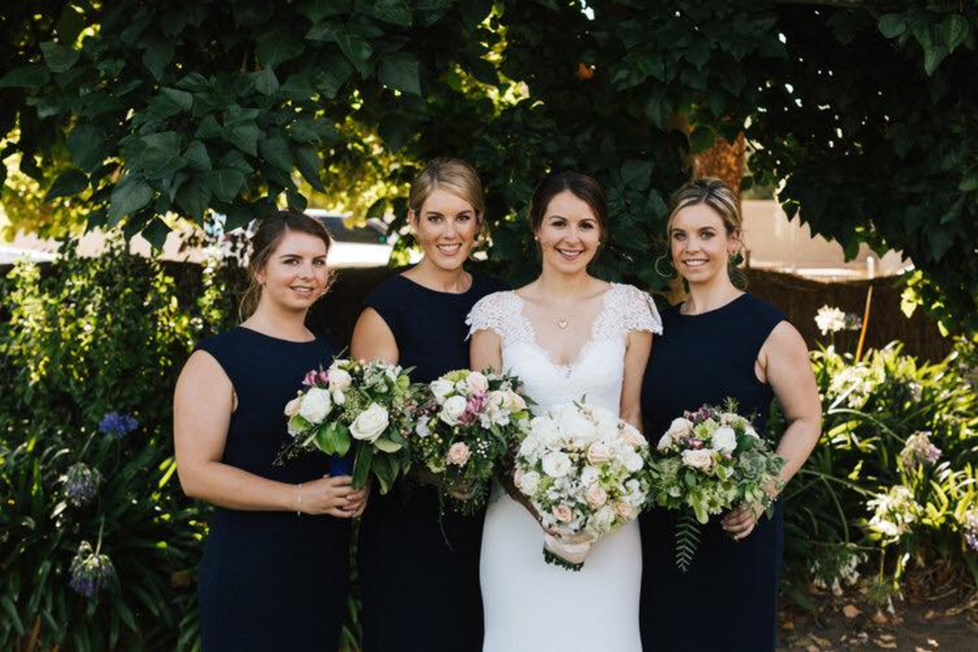 Bride and bridesmaids in navy dresses holding bouquets in a lush garden setting.