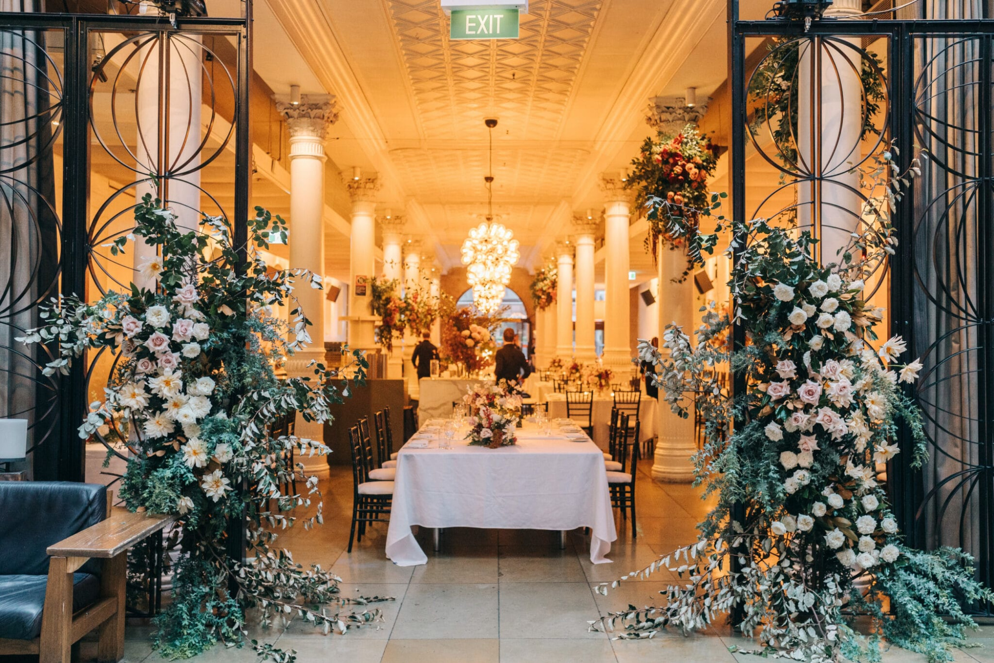 Elegant indoor wedding reception with grand columns, lush floral arrangements, and a long banquet table under warm chandeliers.
