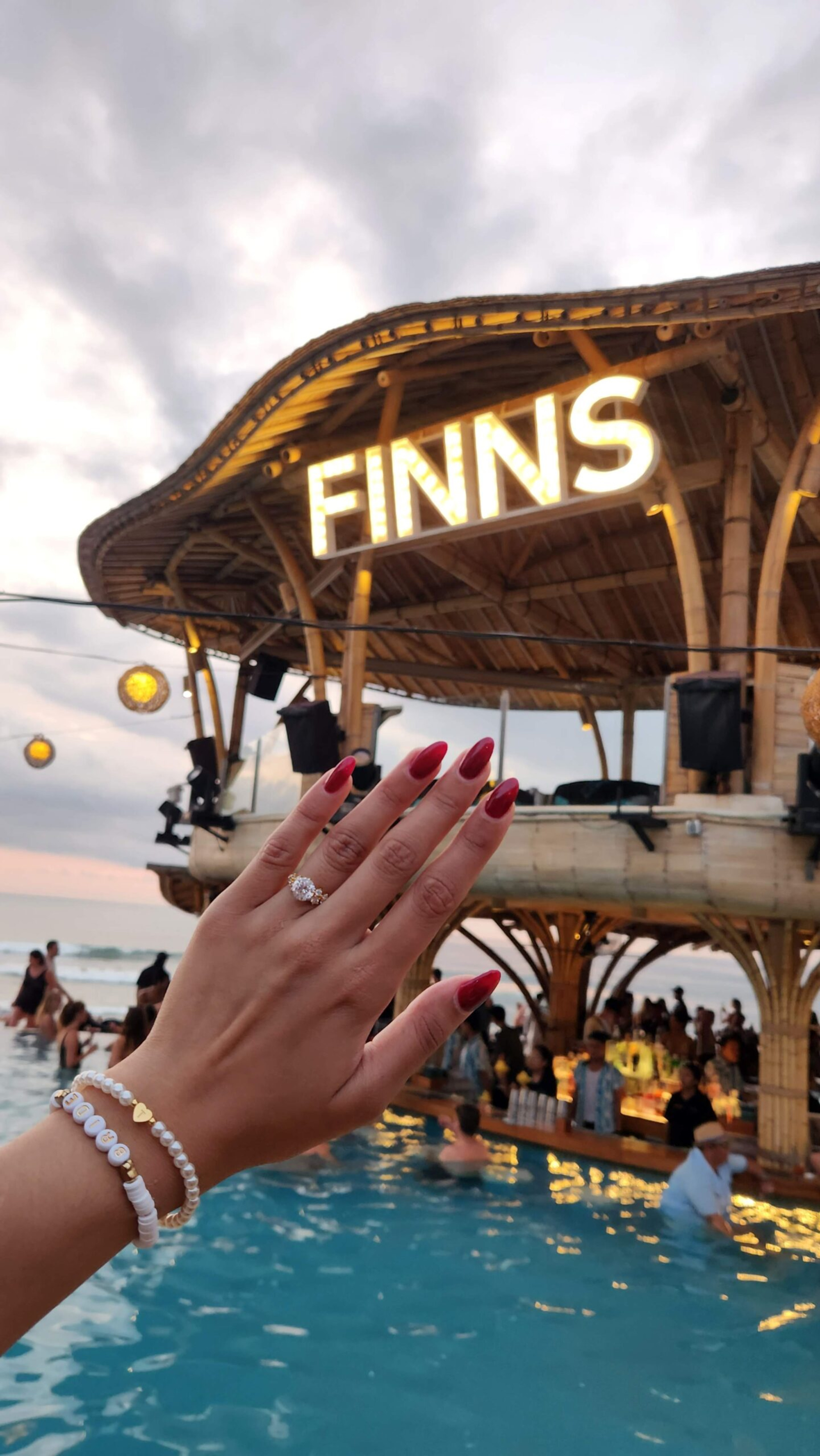 Engagement ring hand posed over a lively pool bar at an oceanfront tropical venue at sunset.