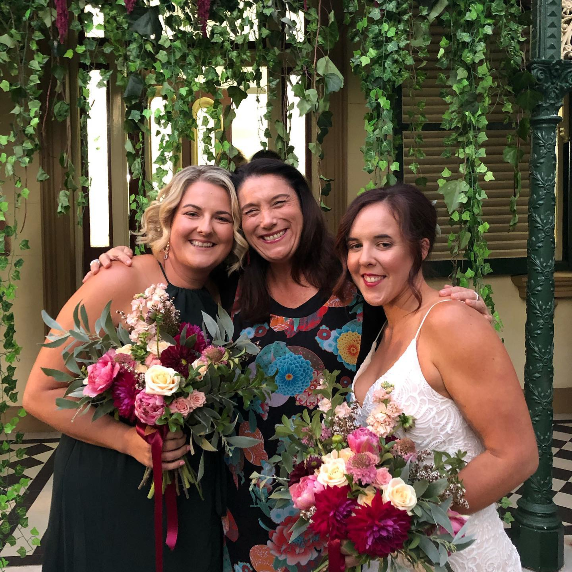 Bride and bridesmaid smiling with vibrant bouquets in a leafy garden setting.