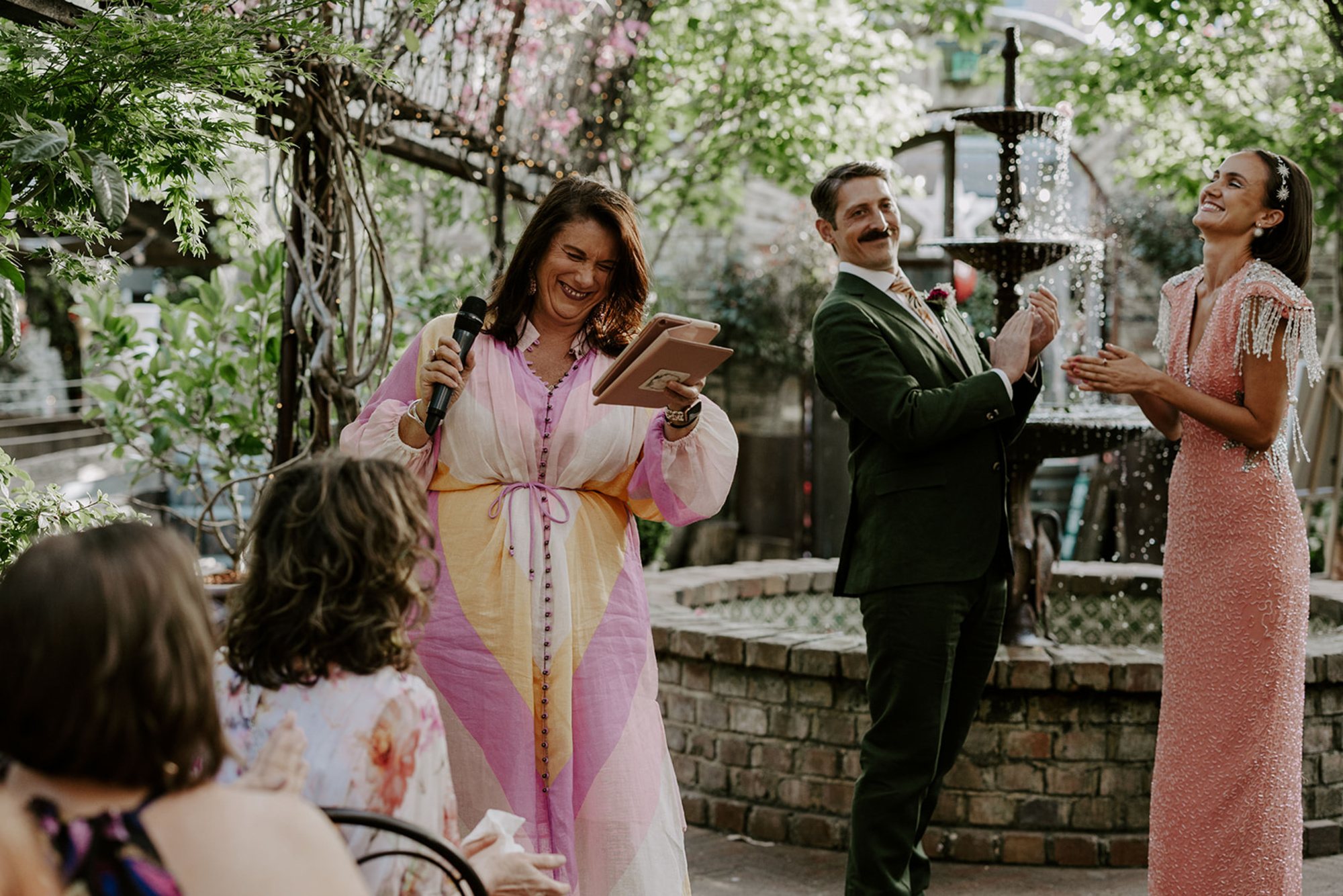 An officiant leads a joyful outdoor wedding ceremony as the couple laughs and applauds by a garden fountain.
