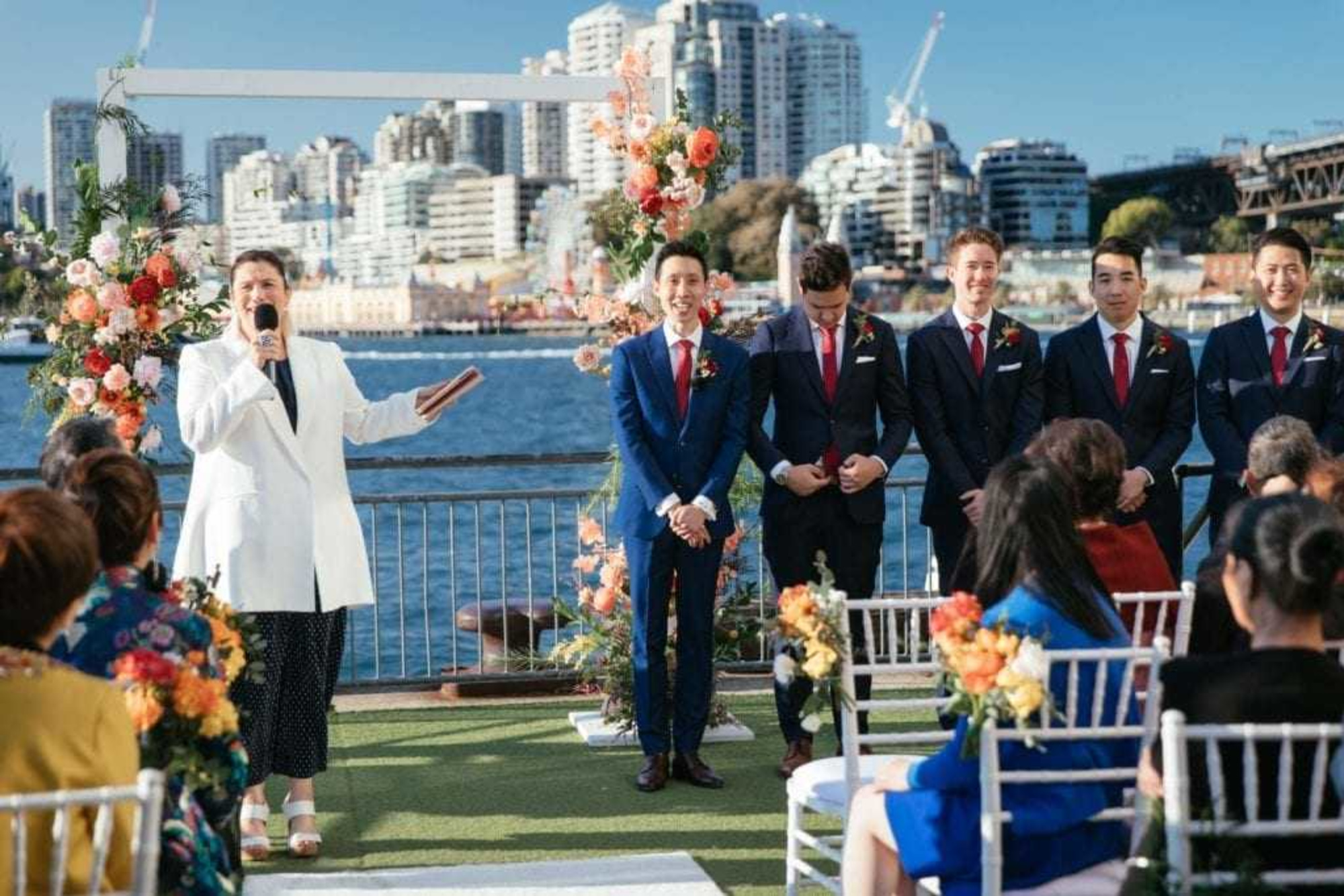 Outdoor waterfront wedding ceremony with officiant, groom, and groomsmen framed by colorful floral arches and city skyline backdrop.