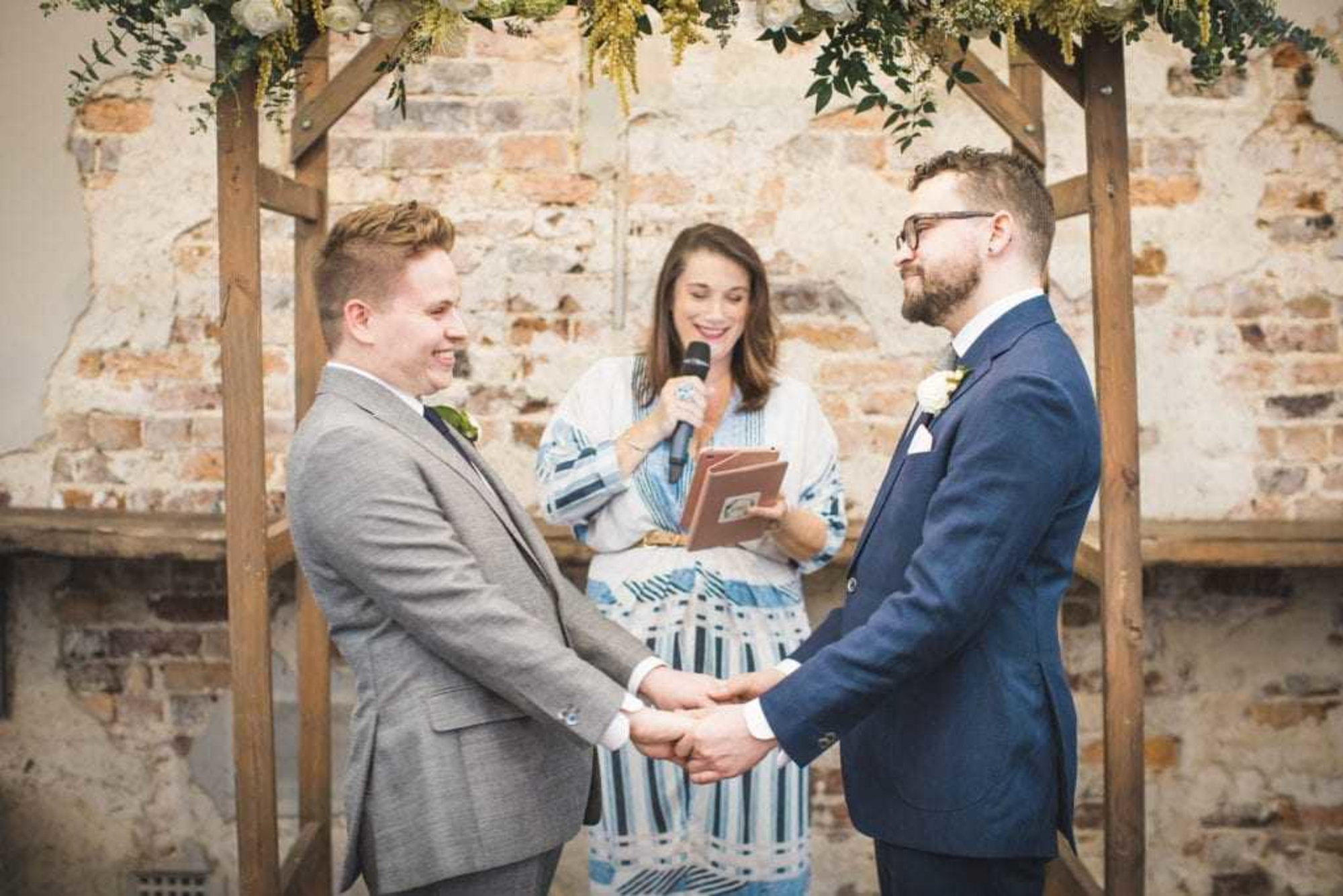 Two grooms hold hands under a rustic floral arch while the officiant reads during their indoor wedding ceremony.