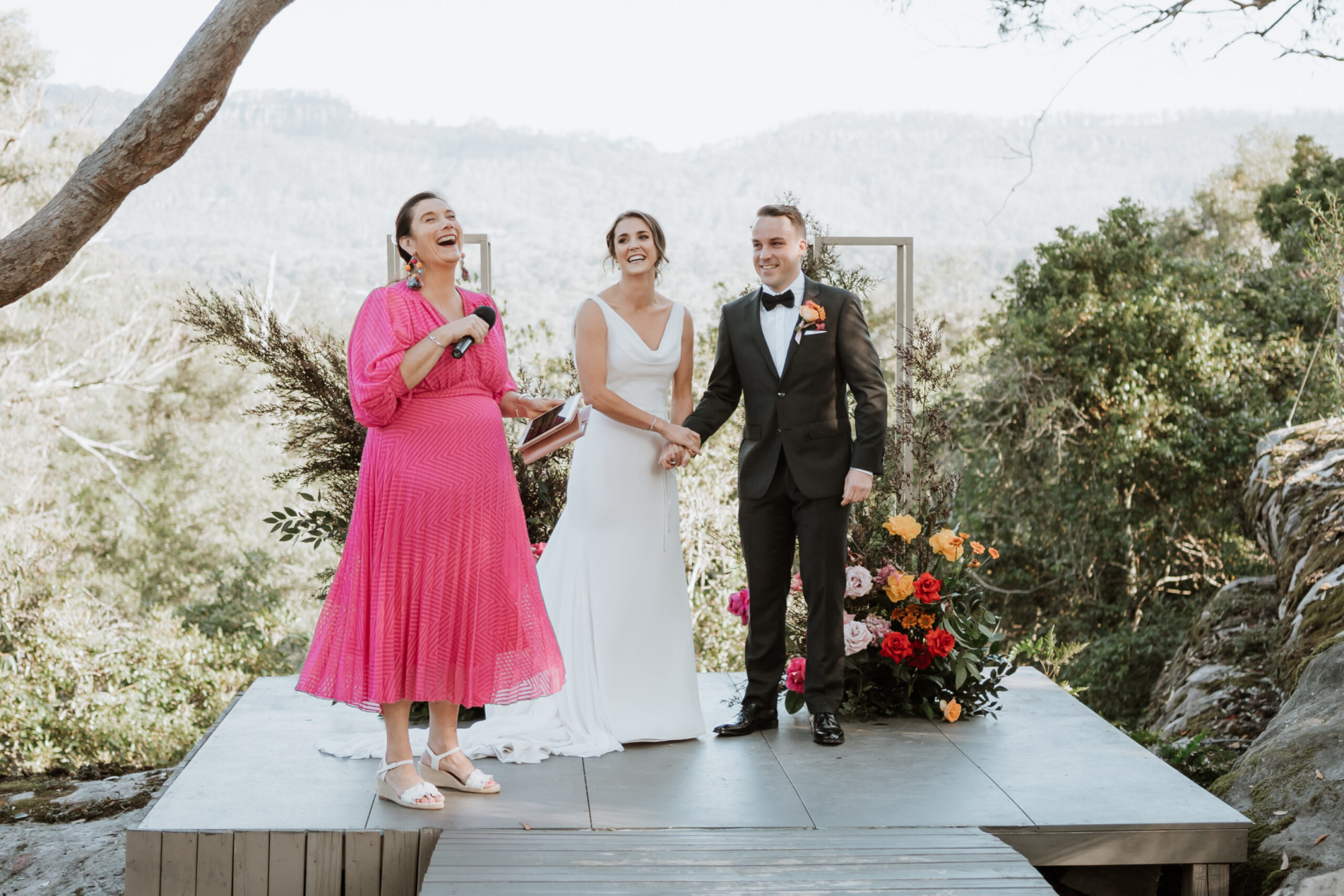 Outdoor wedding ceremony with smiling couple and celebrant on a small platform framed by colorful flowers and mountain views.