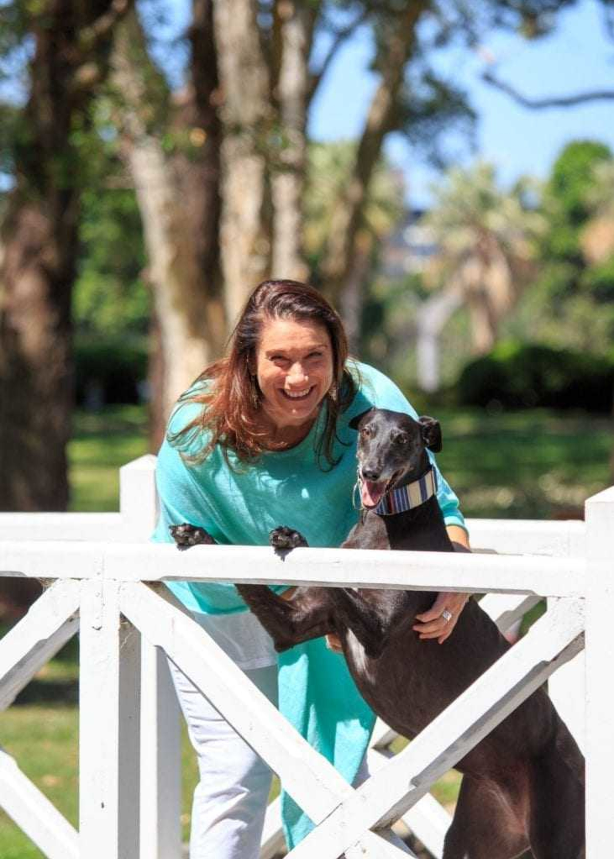 Woman smiling with a black dog at a white fence in a sunny garden setting.