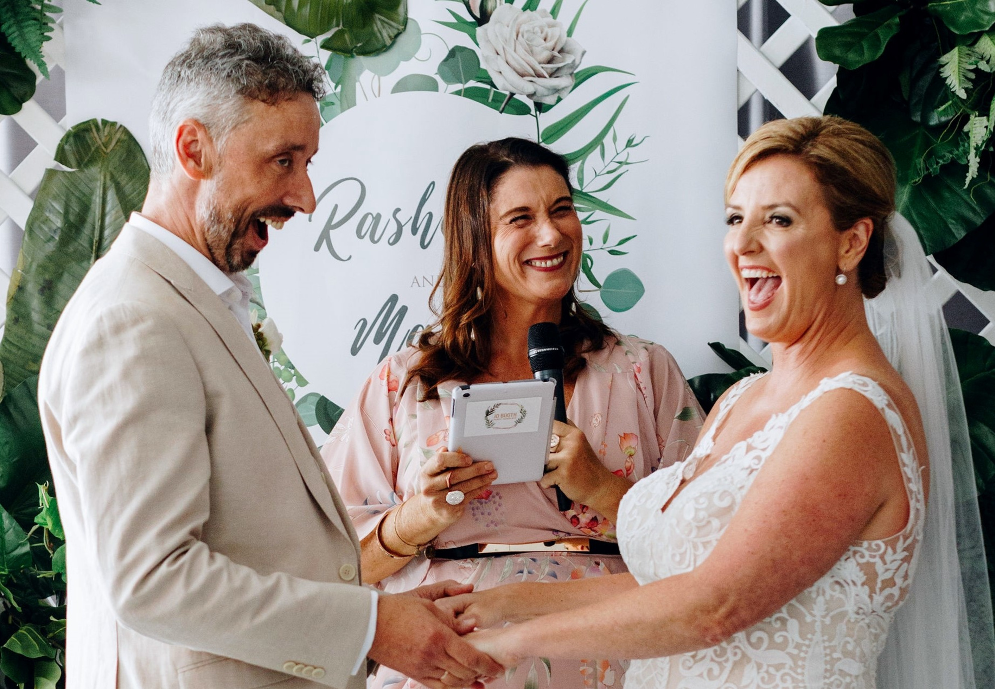 Joyful couple holding hands and laughing with their celebrant during an indoor wedding ceremony.