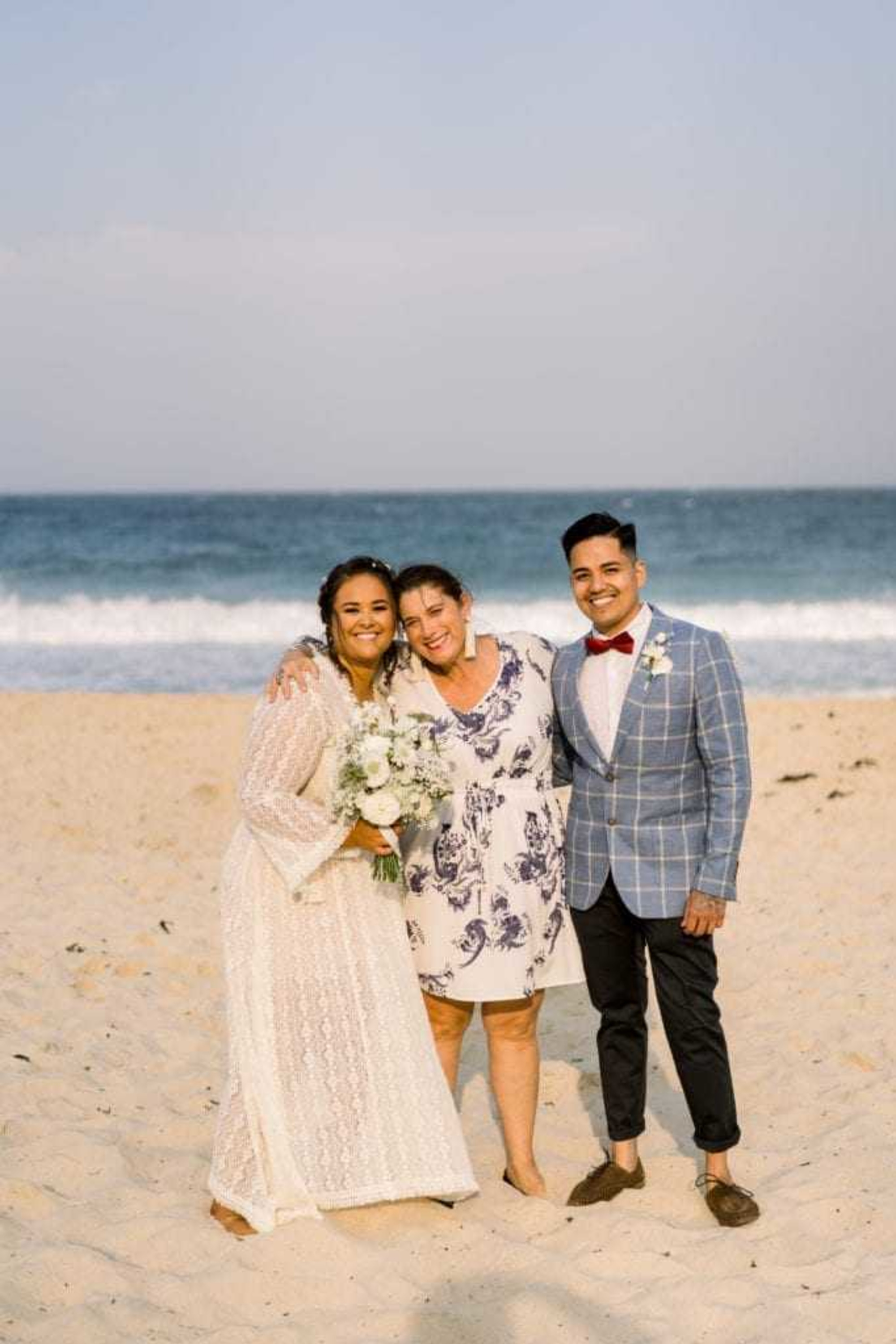 Bride, groom, and officiant smiling together on a sandy beach with ocean waves in the background.