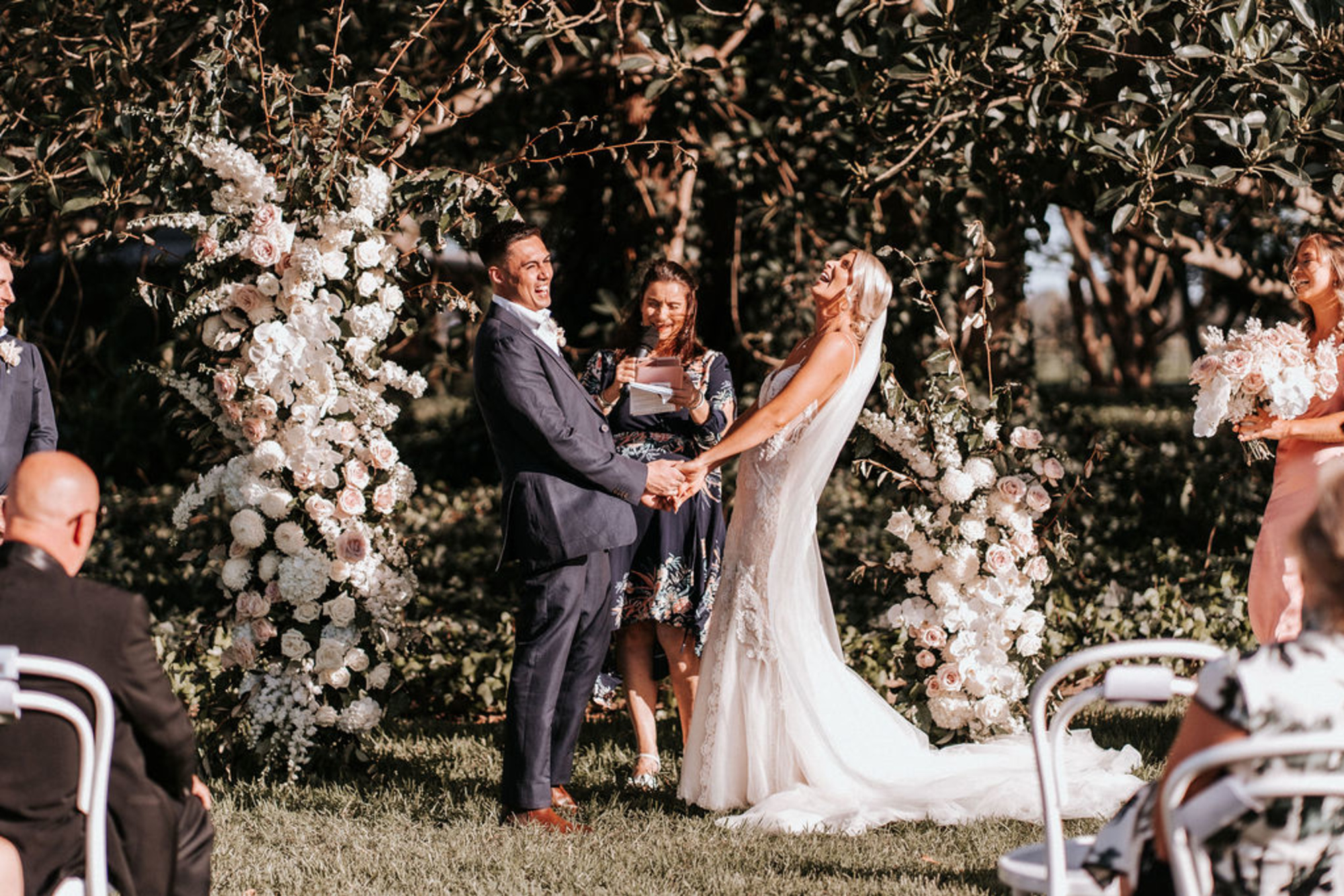 Bride and groom laughing during an outdoor garden ceremony beneath lush white floral arches.