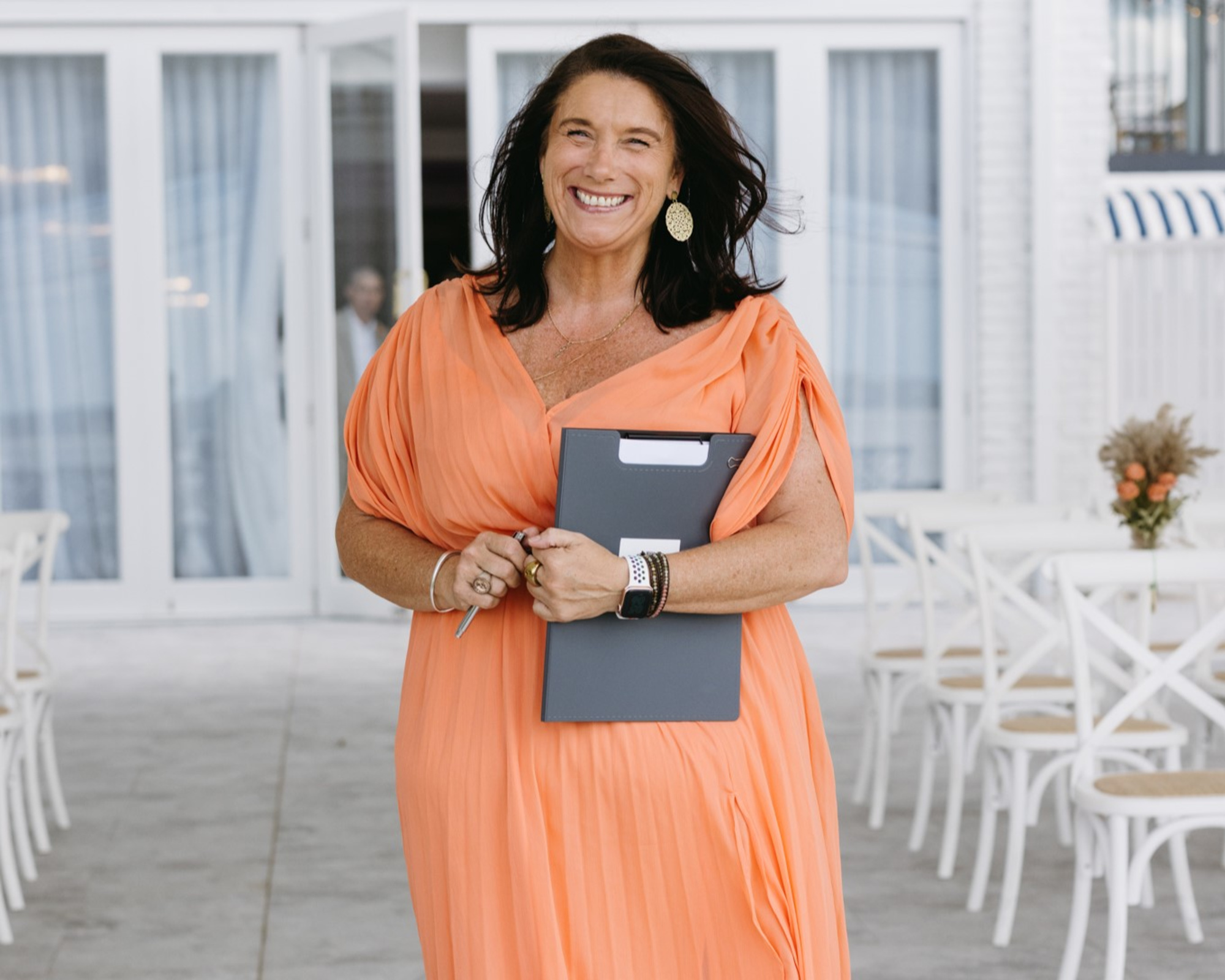 Smiling wedding planner in an orange dress holding a clipboard at a bright outdoor venue with white chairs.