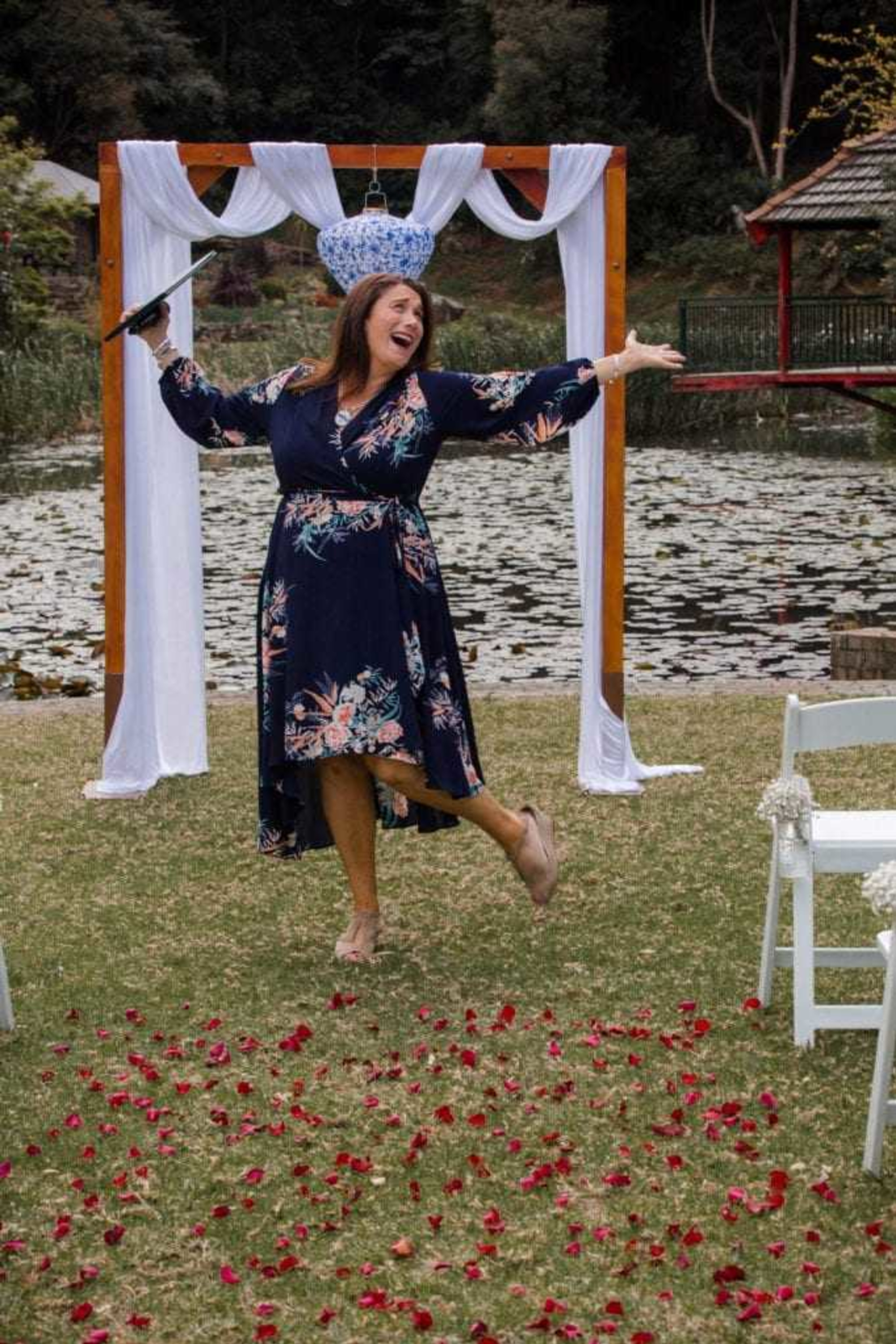 Joyful wedding celebrant posing under a decorated arch by a lakeside aisle scattered with rose petals.